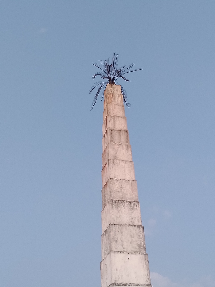A tall, segmented concrete monument, tapering upwards, is depicted against a clear, light blue sky in Ogboko, Nigeria. The structure is light-colored, exhibiting some visible weathering and discoloration. At its apex, a dark, stylized metal sculpture resembling a palm tree with numerous spiky fronds is mounted. No individuals, other structures, or discernible actions are present within the frame.