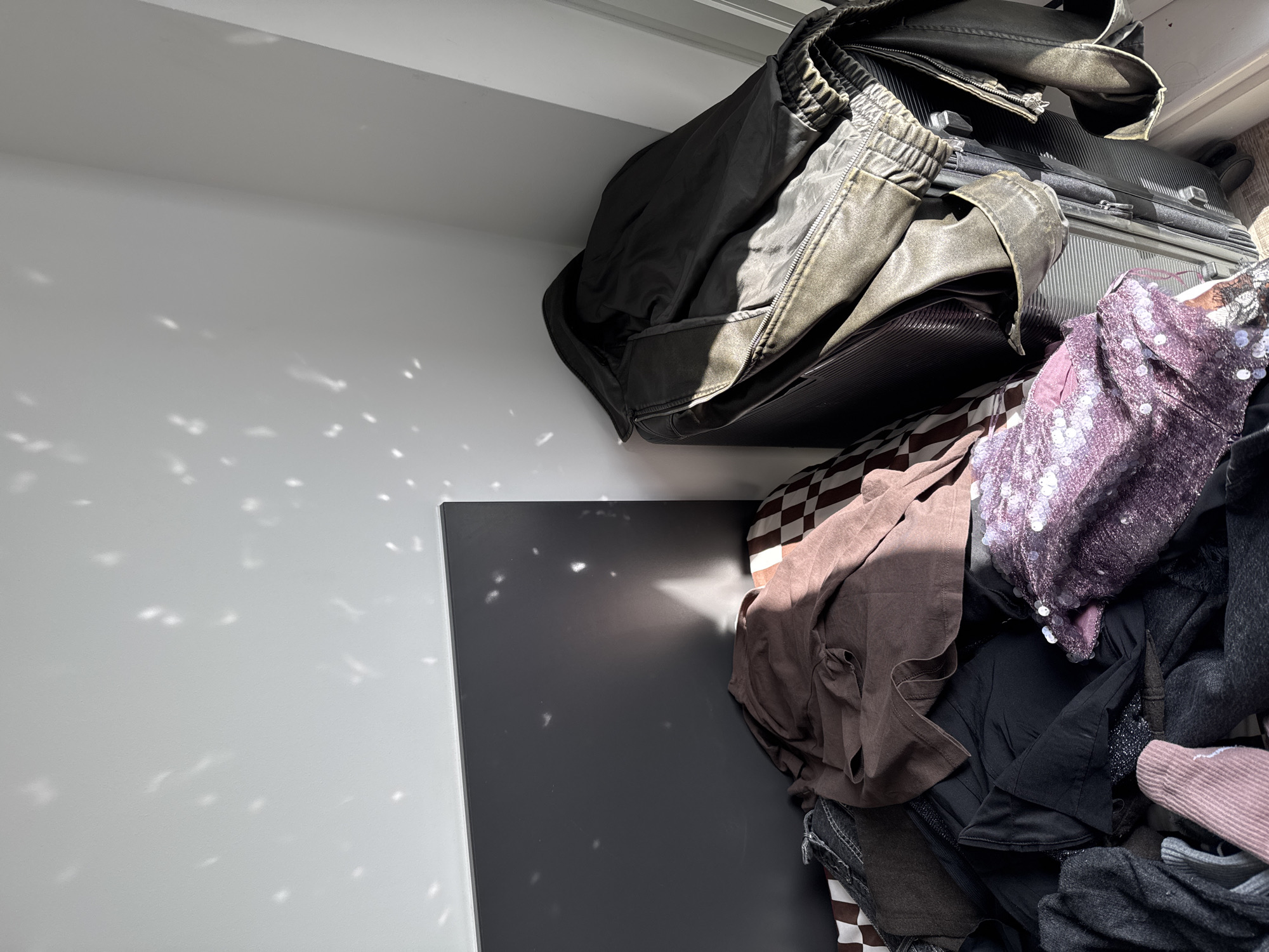 The image shows a cluttered storage space in a location identifiable as Hull, United Kingdom. A black suitcase is partially visible, topped with a dark jacket, suggesting travel or storage of personal items. A pile of clothing in various colors and textures, including a sequined garment, spills out around the suitcase. The storage area is defined by white walls, reflecting dappled sunlight that casts a pattern on the wall and a gray surface, potentially a door or panel.