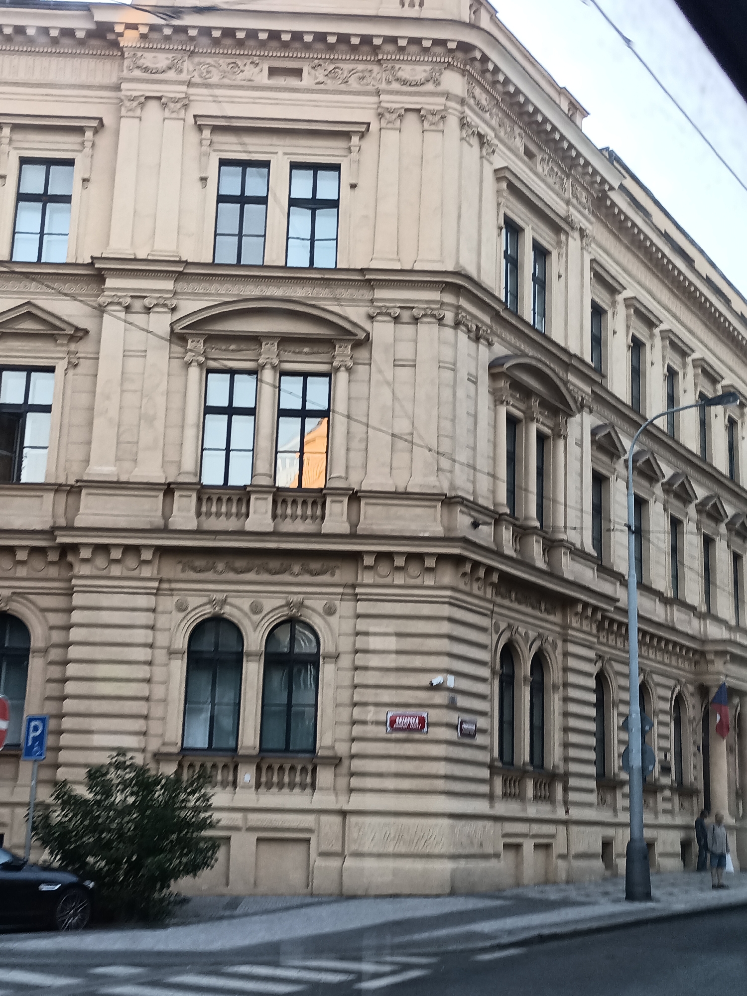 The image shows the facade of a multi-story building in Prague, Czechia. The building has a beige-colored exterior with classical architectural elements, including columns, pediments above windows, and decorative cornices. Numerous windows with dark frames are visible on each level. Some windows reflect the sky or surrounding buildings. A street lamp is positioned in front of the building. A street sign with red and white lettering is mounted on the building's facade. On the sidewalk, a dark-colored car is parked to the left, and a small tree is growing near the building. Two individuals are walking in the distance on the right side of the image, one carrying a white bag. A parking sign is also visible on the left side of the street. The scene appears to be a street view in an urban environment.