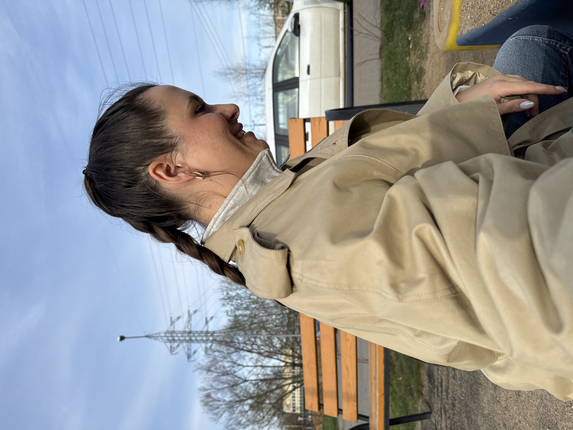 A young woman is sitting on a park bench in Akulovo, Gp Kubinka, Russia. She is wearing a tan trench coat, jeans, and has her hair braided. The woman is smiling and looking to the left. A white van is parked nearby. In the background, there is a utility pole and some trees. The sky is blue with some clouds.