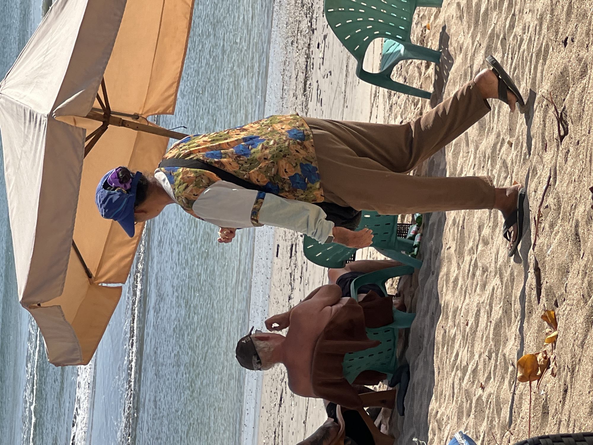 A full shot captures a beach scene in Kuta, Indonesia, under the shade of a large, light-brown beach umbrella. In the foreground, a person wearing a blue cap and a colorful floral shirt walks across the sand, their feet clad in dark flip-flops. To the left, a shirtless man in a brown cap sits on a green beach chair, facing away from the camera, a brown towel draped over his back. The ocean, with gentle waves, forms the background, meeting the sandy shore. Several empty green beach chairs are scattered on the sand. A discarded orange object lies on the sand in the lower left quadrant.