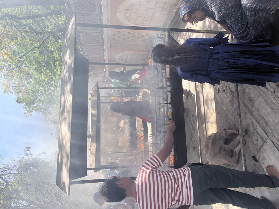 The image shows a group of people standing around a large incense burner. The burner is made of metal and is filled with burning incense sticks. There is a lot of smoke in the air. The scene is set in Panchkula, India, at the coordinates 30.722, 76.855. There is a young boy in a striped shirt facing the camera, and he is holding a small, burning object over the fire. A woman in a blue dress is standing on the right, looking down at the burning incense. An older woman in a floral patterned headscarf is seated on the right, and looking up at the camera. A dog is sleeping in the foreground. The scene is in a partially enclosed area with stone walls and a stone walkway. Green foliage can be seen through the open areas.
