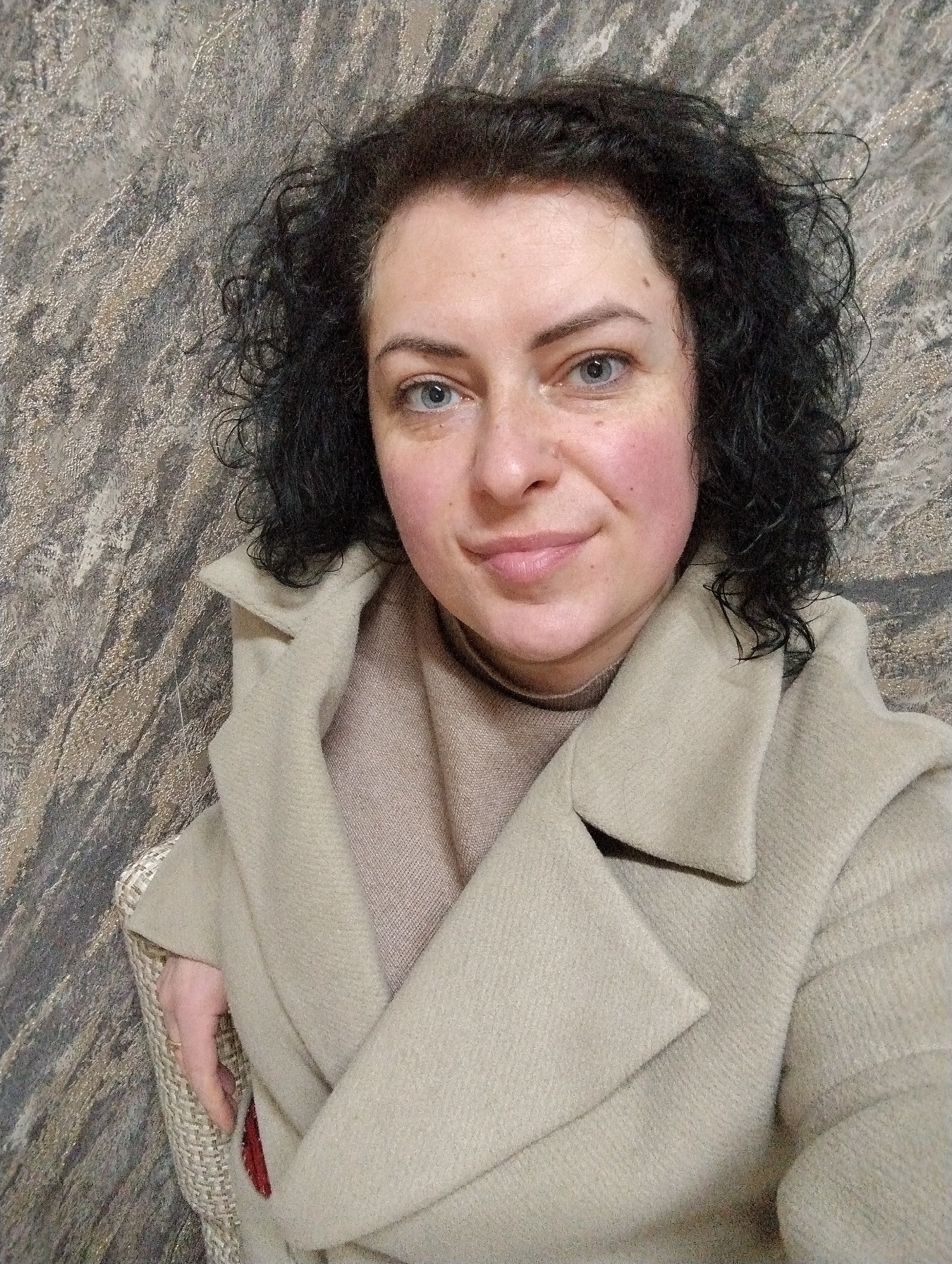 A woman with fair skin and dark, curly hair is centered in the image. She has blue eyes, and her cheeks appear slightly flushed. She is wearing a light beige coat over a beige turtleneck sweater. The coat has a visible collar and lapel. Her left hand is resting on the arm of a chair, which has a light-colored woven texture. The background is a mottled gray and beige pattern. There is no verifiable indication of the location being Grishanki, Russia. The scene appears to be indoors.