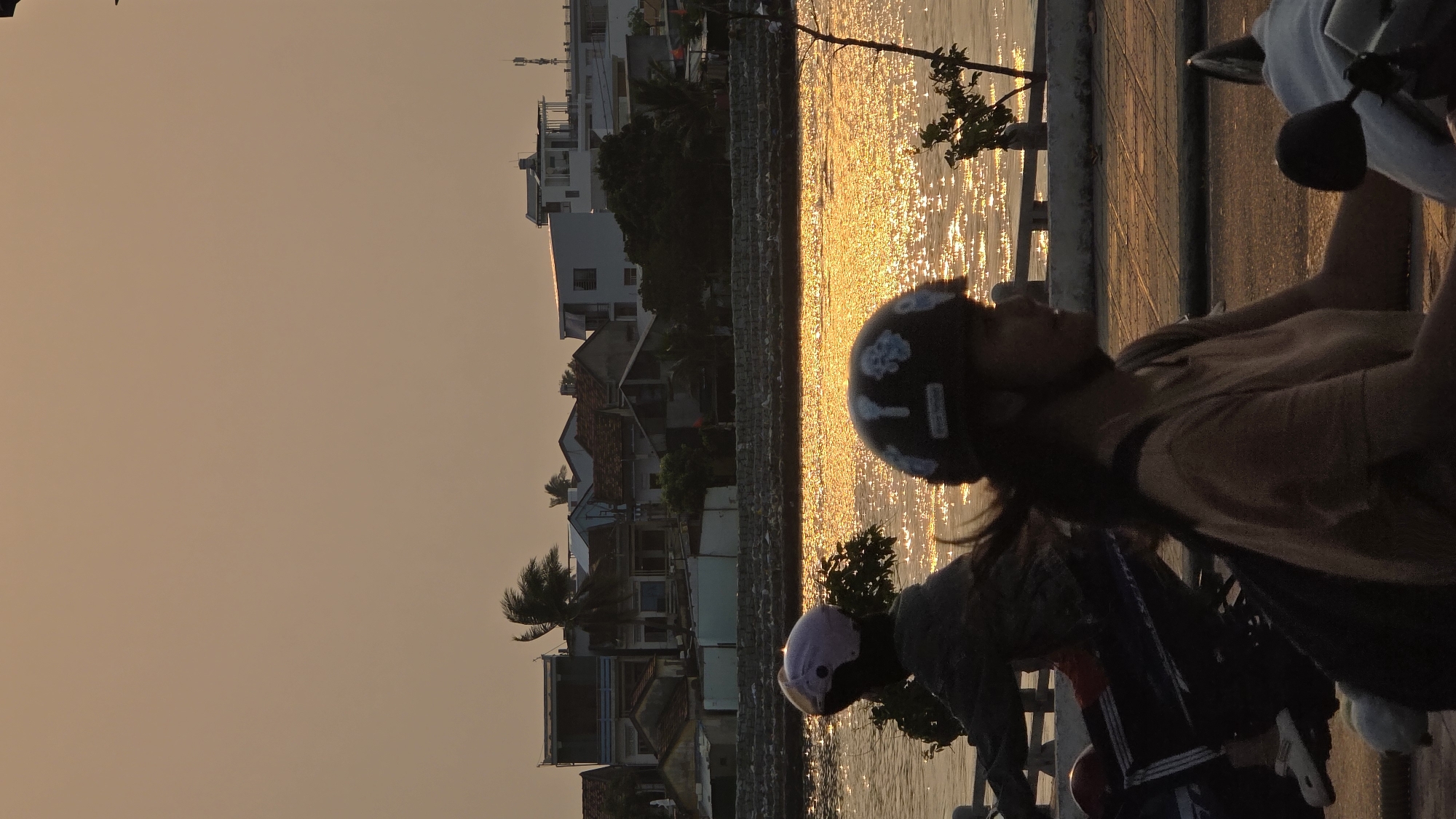 Two people are riding motorbikes along a paved path in Nha Trang, Vietnam, at sunset. The golden light reflects off the water next to the path, which is lined with a low concrete wall. In the background are buildings and trees. The first person is wearing a dark helmet with white decorations and a light-colored shirt. The second person is wearing a light pink helmet and a dark jacket.