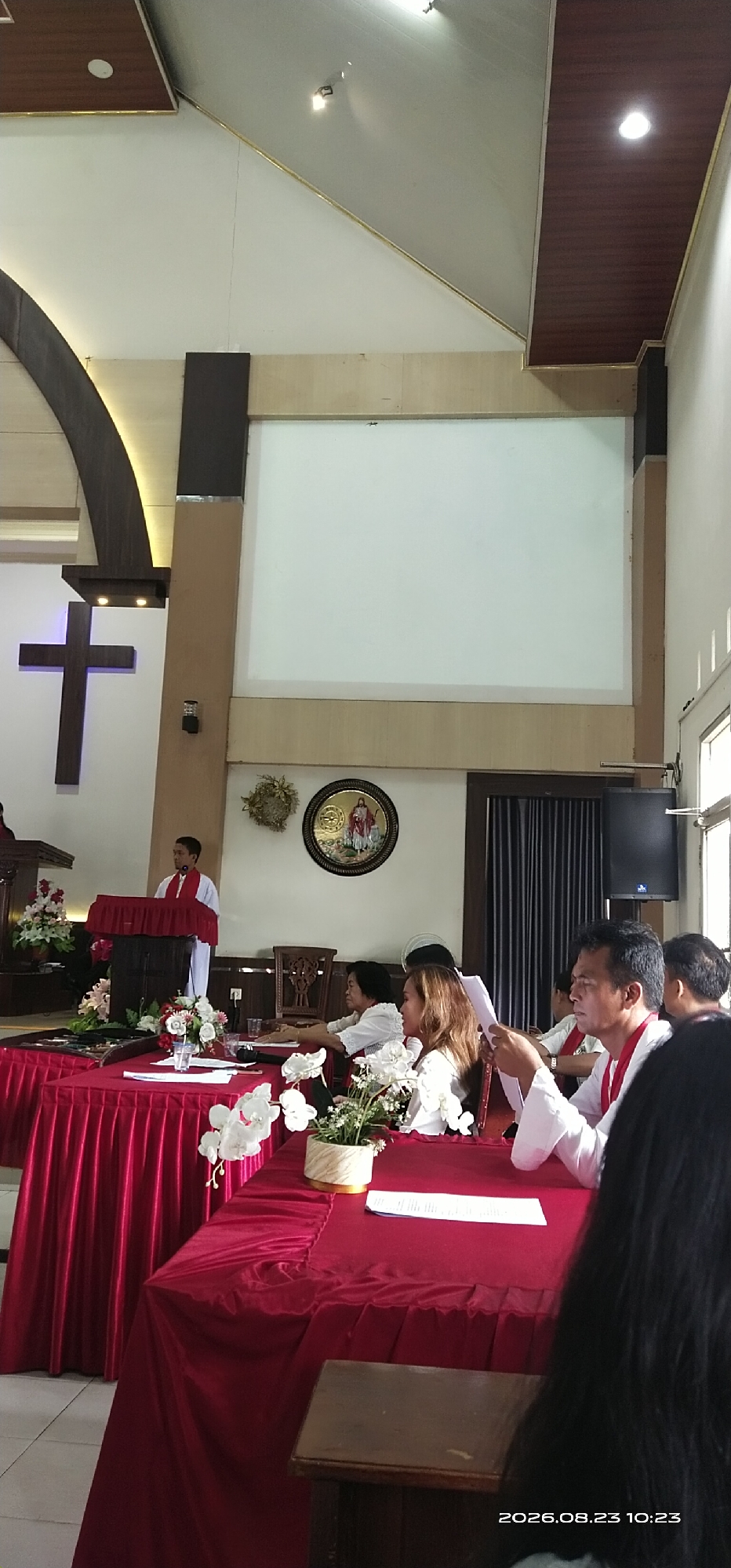 This image captures an indoor scene that appears to be a church or religious hall. The room is decorated with red draped tablecloths on several tables. A large dark brown cross is mounted on the left wall. In the center of the room, a man in white clerical attire is standing at a podium, likely delivering a speech or leading a service. Several individuals are seated at the tables, some appearing to be listening to the speaker, while others are engaged in reading documents. The background features a large blank screen, decorative wall hangings, and dark curtains. The lighting suggests an artificial illumination source. The timestamp "2026.08.23 10:23" is visible in the lower right corner. Based on the provided information, this scene is located in the city of Jaar, Indonesia.