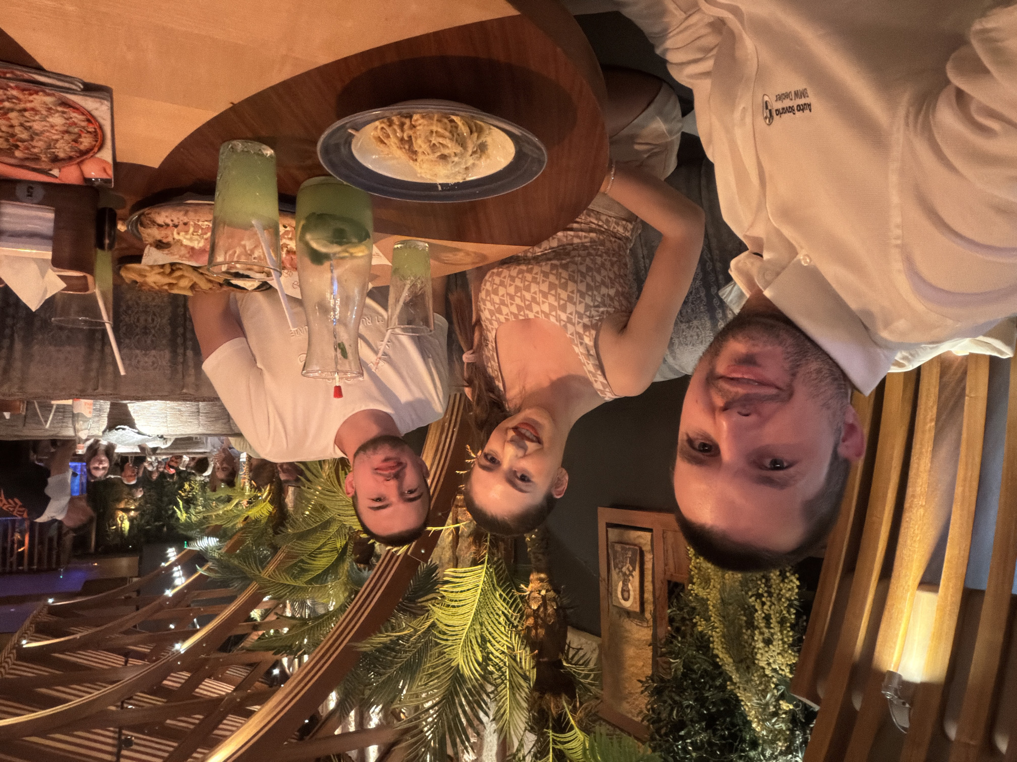 The image depicts three individuals – two males and one female – seated at a circular wooden table within a restaurant, likely a branch of **Happy Bar & Grill** in **Sofia, Bulgaria**.
The man on the left, foreground, wears a white collared button-down shirt featuring an embroidered "Auto Bavaria BMW Dealer" logo on the left chest. He has short dark hair and light stubble, smiling directly at the camera. To his right, the woman wears a light-colored patterned tank top and smiles towards the camera, with her dark hair partially pulled back. To her right, the second man wears a white short-sleeved t-shirt, has short dark hair and light facial hair, looking towards the camera with a slight smile.
The table holds a variety of food and drinks. In the foreground, a white plate with a blue rim contains a serving of pasta topped with grated cheese. A tall glass pitcher filled with green liquid, lemon slices, and mint is positioned in the center. Two clear glasses with green liquid and white straws are also on the table. Further back, a plate of french fries and a pizza are visible. A brown wooden napkin holder with tissues and a menu displaying a pizza are also present. A partially obscured water bottle is also on the table.
The restaurant's interior features elaborate wooden slatted structures forming arches and panels on the ceiling and walls, giving it an open yet enclosed feel. Lush green tropical plants, including palm trees and hanging foliage, are integrated throughout the decor. Warm, dim artificial lighting emanates from fixtures within the wooden structures. In the background, other patrons are visible at their tables, and a staff member in a black t-shirt is partially seen on the far right. A framed Eastern Orthodox icon is visible on a dark wall section to the left, behind the woman.