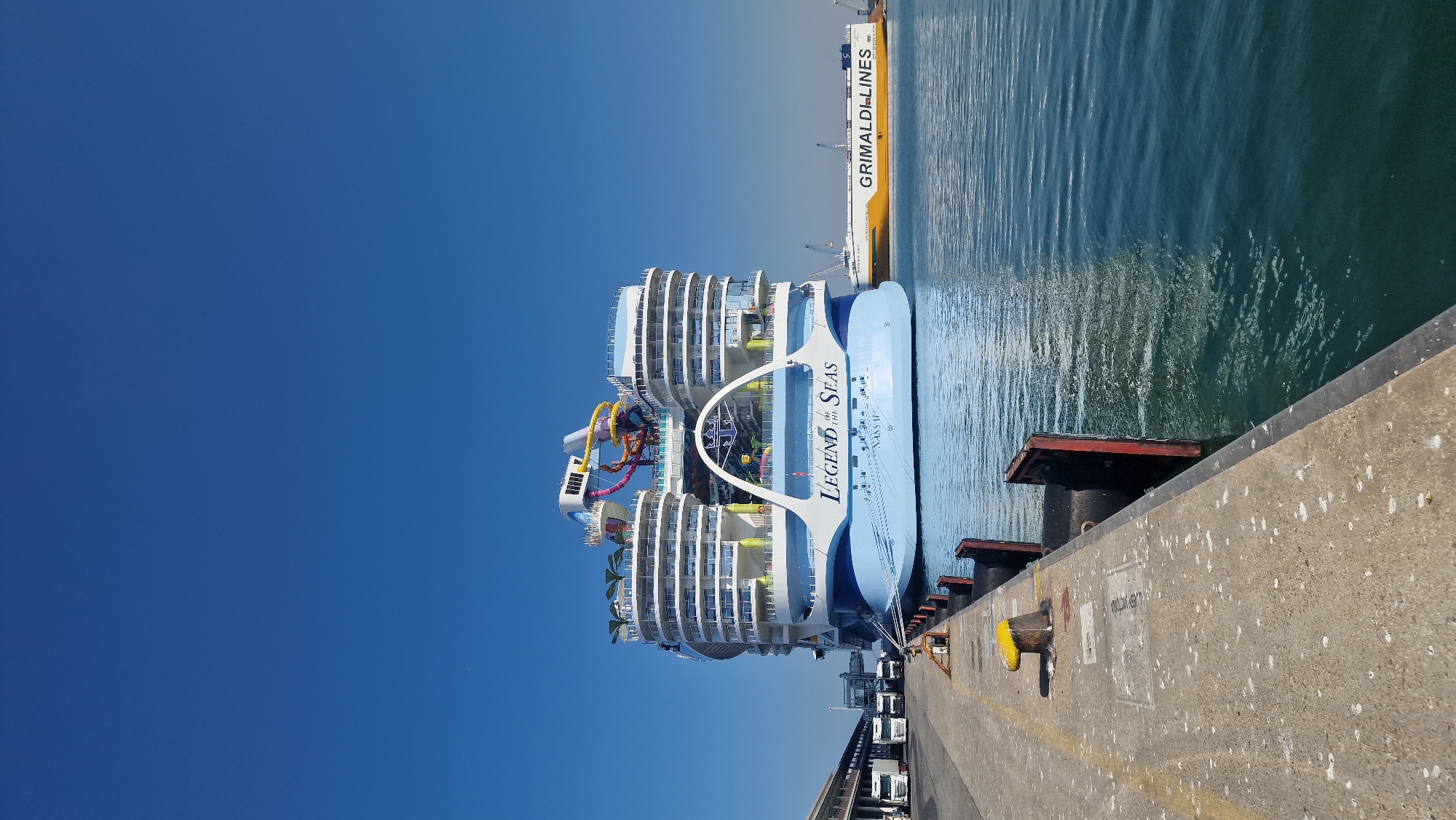 A large cruise ship, the "Legend of the Seas," is docked at the port of Civitavecchia, Italy, on a clear, sunny day. The ship, predominantly white and light blue, features multiple decks with balconies and a distinctive amusement area with colorful water slides at the top. The ship's name and "Nassau" are visible on its hull. To the right of the cruise ship, a yellow and white cargo ship with "GRIMALDI LINES" written on its side is also present. In the foreground, a concrete pier runs along the left side of the frame, with yellow bollards, black fenders, and several white trucks parked along its edge. The water is calm and reflects the blue sky and the ships. The overall scene depicts a bustling port environment with maritime vessels.