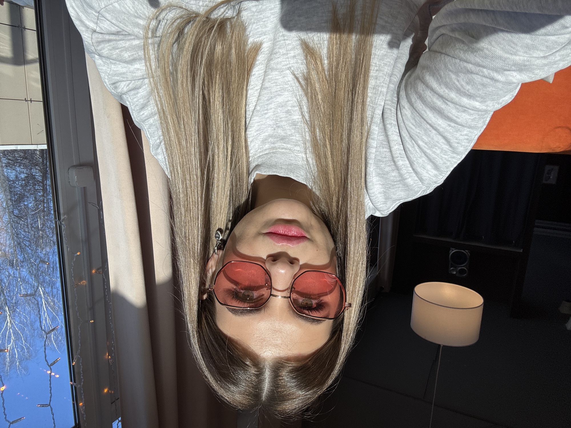 The image presents a head-and-shoulders selfie of a young adult female in an indoor setting, identified as being in the city of Chaikovsky, Russia. The subject is centered, looking directly at the camera. She has long, straight light brown hair parted in the middle. She wears rose-tinted, octagonal-framed glasses with thin metallic frames, pink lipstick, and small silver-toned heart-shaped drop earrings. Her attire consists of a light gray, ribbed, high-necked, long-sleeved top.
Strong natural sunlight enters from the right side of the frame, illuminating the subject's right side and casting distinct shadows on her face and left arm. The background to the subject's left is a dimly lit interior space featuring a white, drum-shaped pendant light fixture suspended from a dark ceiling. Below the lamp, a dark object resembling an audio speaker with a circular driver is visible. Part of an orange upholstered surface, likely a sofa or chair, is visible behind her left shoulder.
To the subject's right, a window reveals an outdoor scene under a clear, bright blue sky. Bare deciduous trees are visible against the sky, indicating a late autumn or winter season. A light-colored building with rectangular facade panels is seen through the window. Inside the window frame, a string of decorative lights with small, warm-toned bulbs hangs vertically. Light-colored curtains are partially drawn on the indoor side of the window.