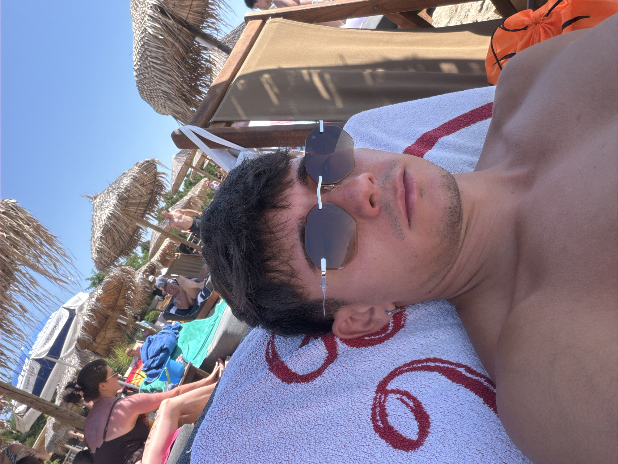 A young man with dark hair and sunglasses is lying on a towel on a beach lounge chair. He is wearing sunglasses and has a silver hoop earring in his left ear. The towel has a large red circular design on it. In the background, there are thatched umbrellas, other people relaxing on the beach, and the blue sky. The scene appears to be in the city of Thassos, Greece, likely at a beach resort.