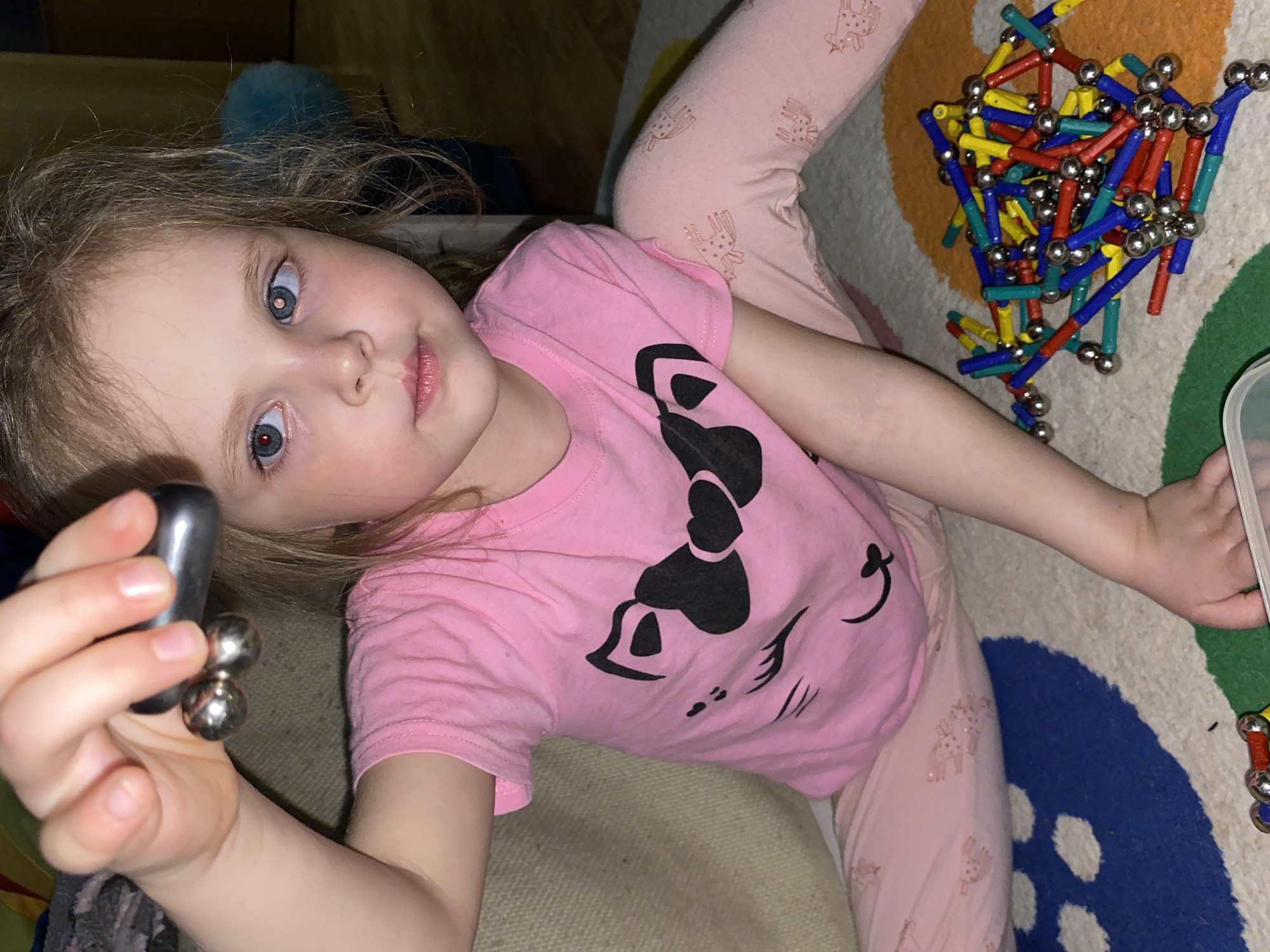 A young girl, with light brown hair and wide blue eyes, is captured in a close-up, high-angle shot, looking directly at the viewer. She appears to be lying down or reclining on a light-colored surface, possibly a cushion or blanket, next to a colorful, patterned carpet. Her expression is calm and neutral, with a slight pout to her lips.
She is wearing a pink short-sleeved t-shirt featuring a large black graphic of what appears to be a stylized cat face or a similar animal design, and light pink pajama bottoms adorned with small, repetitive patterns of what might be reindeer or deer.
In her left hand, she holds two small, silver magnetic spheres attached to a darker, cylindrical magnetic bar. To her right, scattered on the patterned carpet, is a pile of colorful magnetic construction toys, comprising numerous red, yellow, blue, and green plastic sticks connected by silver magnetic balls, suggesting she was actively playing. The carpet itself displays abstract shapes in hues of blue, green, orange, and white.
The setting is indoors, likely a home or play area, illuminated by what appears to be artificial light, possibly from a camera flash, given the reflections in her pupils. There is no visible text in the image. This scene takes place in Votkinsk, Russia.