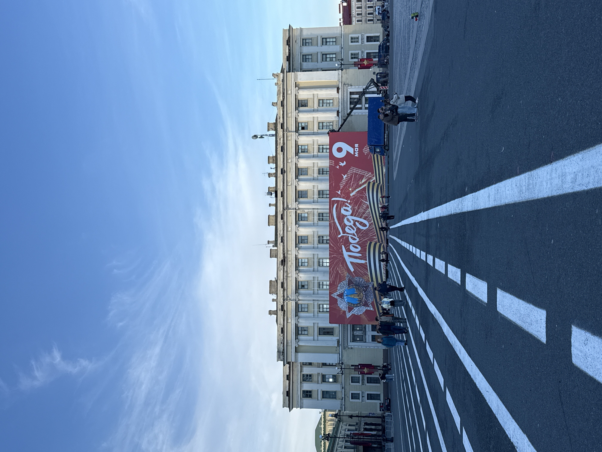 A vertical medium shot shows Victory Day in Saint Petersburg, Russia. The sky is clear, light blue with wispy white clouds. A large beige building with white pillars lines the street. In front of the building is a large red banner with "Victory!" written in Russian and the date "May 9". Several people stand along the street and in front of the banner. White lines are painted on the street, dividing it into lanes. A truck with a crane is visible behind the banner.