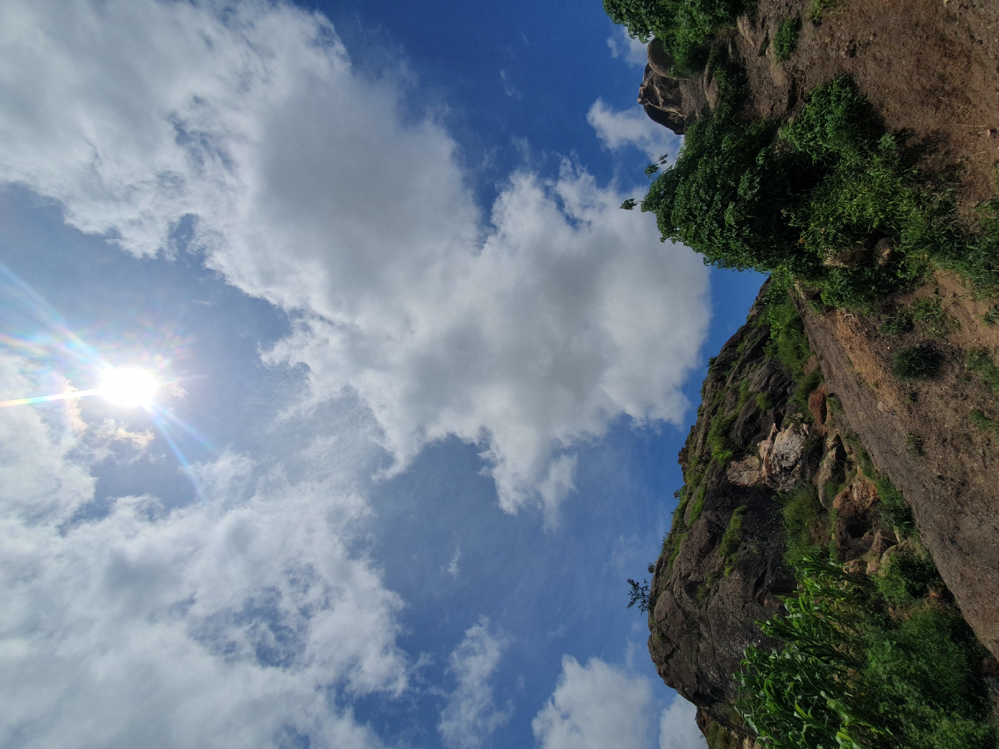 The image captures a low-angle view of a rocky hillside under a bright, partially clouded sky. The sun, high in the sky, creates a lens flare effect with visible rainbow streaks. White, fluffy cumulus clouds are scattered across a vibrant blue sky. The hillside is covered in rough, brown earth and rocks, with patches of green vegetation and bushes. A few larger rocks and small trees are visible on the slope. In the foreground to the left, there are tall green plants that resemble corn stalks. To the right, a dense cluster of green foliage grows on the side of the hill. The overall impression is of a natural, somewhat rugged landscape. The location is Gura Top, Nigeria.
