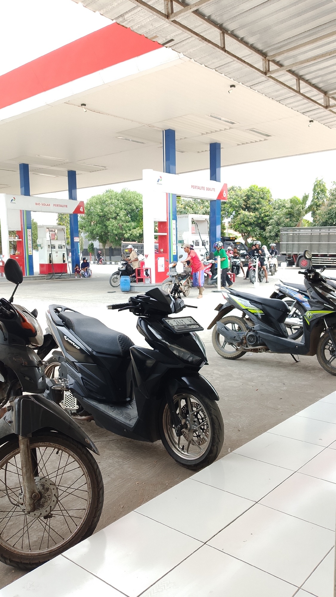 The image depicts a busy outdoor scene at a gas station, identified as being in Kalikangkung, Indonesia. The foreground is dominated by the front wheel and lower body of a black scooter, partially obscuring another scooter to its left. In the midground, several more motorcycles are parked or in use at the gas pumps. A black scooter with a license plate reading "G 2614 HG" is prominently visible. Several people are interacting at the gas pumps. One individual in a red shirt and blue shorts is attending to a motorcycle. Another person in a green shirt and helmet is also near a pump. In the background, the gas station's infrastructure is visible, including white and red structures with blue columns, and signage indicating "PERTALITE DEXTLITE" and "BIO SOLAR PERTALITE". A large truck is parked further in the background. Trees and vegetation surround the gas station, suggesting an open, possibly suburban or rural setting. The ground is paved with concrete and white tiles in the foreground. The overall atmosphere appears to be one of daily activity at a service station.