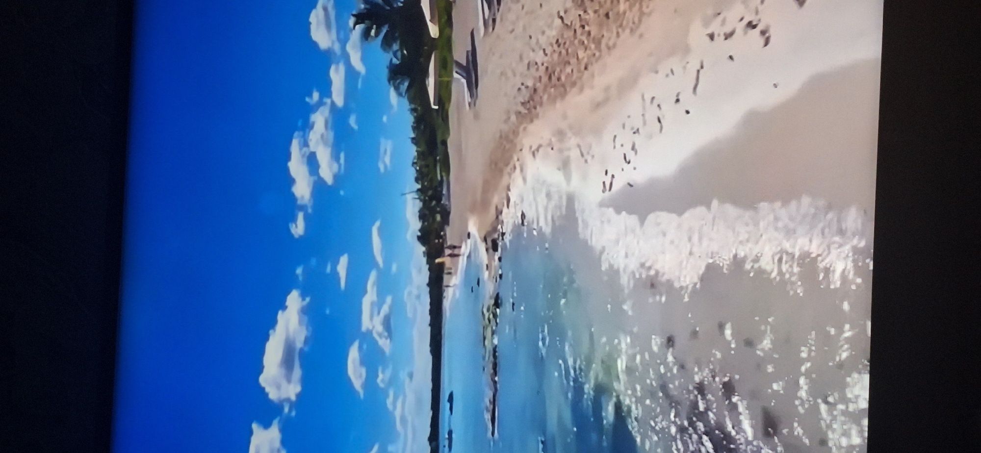 The image depicts a tranquil beach scene under a clear blue sky dotted with fluffy white clouds. The foreground shows gentle waves lapping at a sandy shore, with some dark rocks visible in the shallow water. Further down the beach, several figures are walking, and loungers with umbrellas are set up. In the background, lush green trees, possibly palm trees, line the coast. The scene evokes a sense of peace and relaxation, consistent with a tropical or subtropical vacation destination. There is no indication that this location is in Bila Tserkva, Ukraine, as the environment and vegetation do not align with the typical landscape of that region.