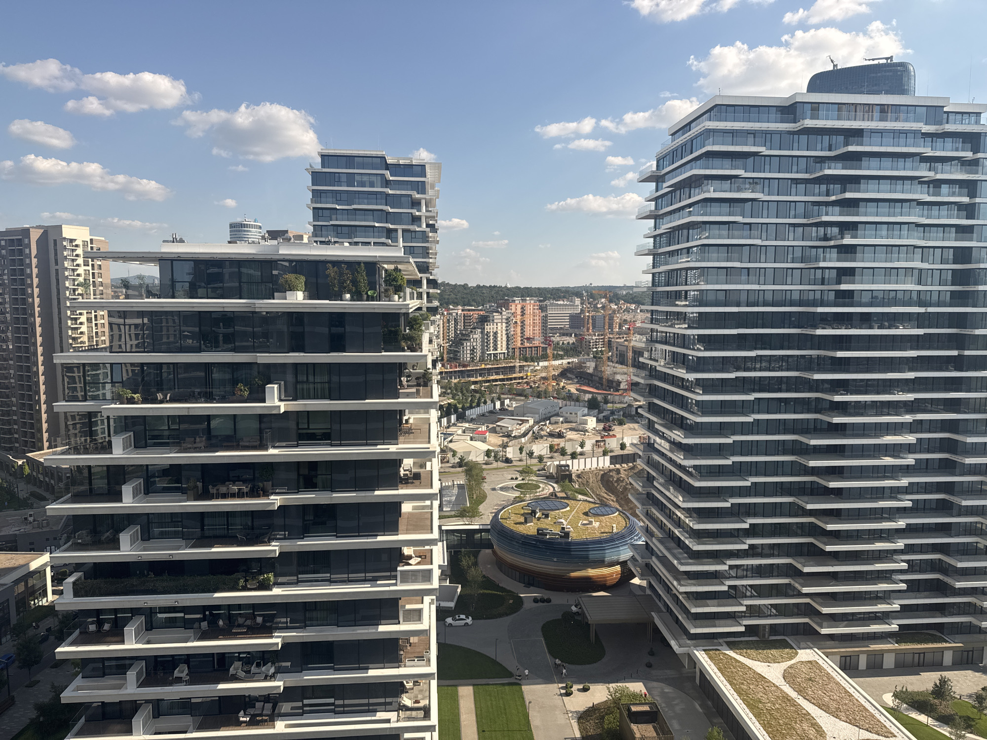 This is a wide-angle, high-angle shot of a modern urban development under a bright blue sky with scattered white clouds. The scene is dominated by several contemporary high-rise buildings with extensive glass facades and layered balconies. Two prominent apartment buildings, one on the left and one on the right, are the main subjects, showcasing their tiered architectural design.
In the middle ground, a distinctive, circular building with a green, undulating roof and colorful banded walls is visible, suggesting a community center or commercial space. Beyond these, a sprawling cityscape of Belgrade, Serbia, unfolds, characterized by a mix of taller residential towers, lower buildings, and construction cranes, indicating ongoing development. The surrounding landscape includes rolling hills and lush greenery, typical of the Belgrade region.
The time of day appears to be late morning or early afternoon, given the bright sunlight casting clear shadows and the clarity of the sky. The weather is fair and sunny, conducive to outdoor activities, though the image focuses on the architecture and urban landscape rather than people engaged in such activities.
Notable details include the precise lines of the buildings, the reflective quality of the glass, and the small plants and outdoor furniture visible on some of the balconies. The presence of a white car on a paved path near the circular building and a few figures walking on a pathway in the foreground suggest a normal day in this residential and commercial complex. The scene conveys a sense of modern urban living and ongoing growth in Belgrade. There is no visible text in the image.
