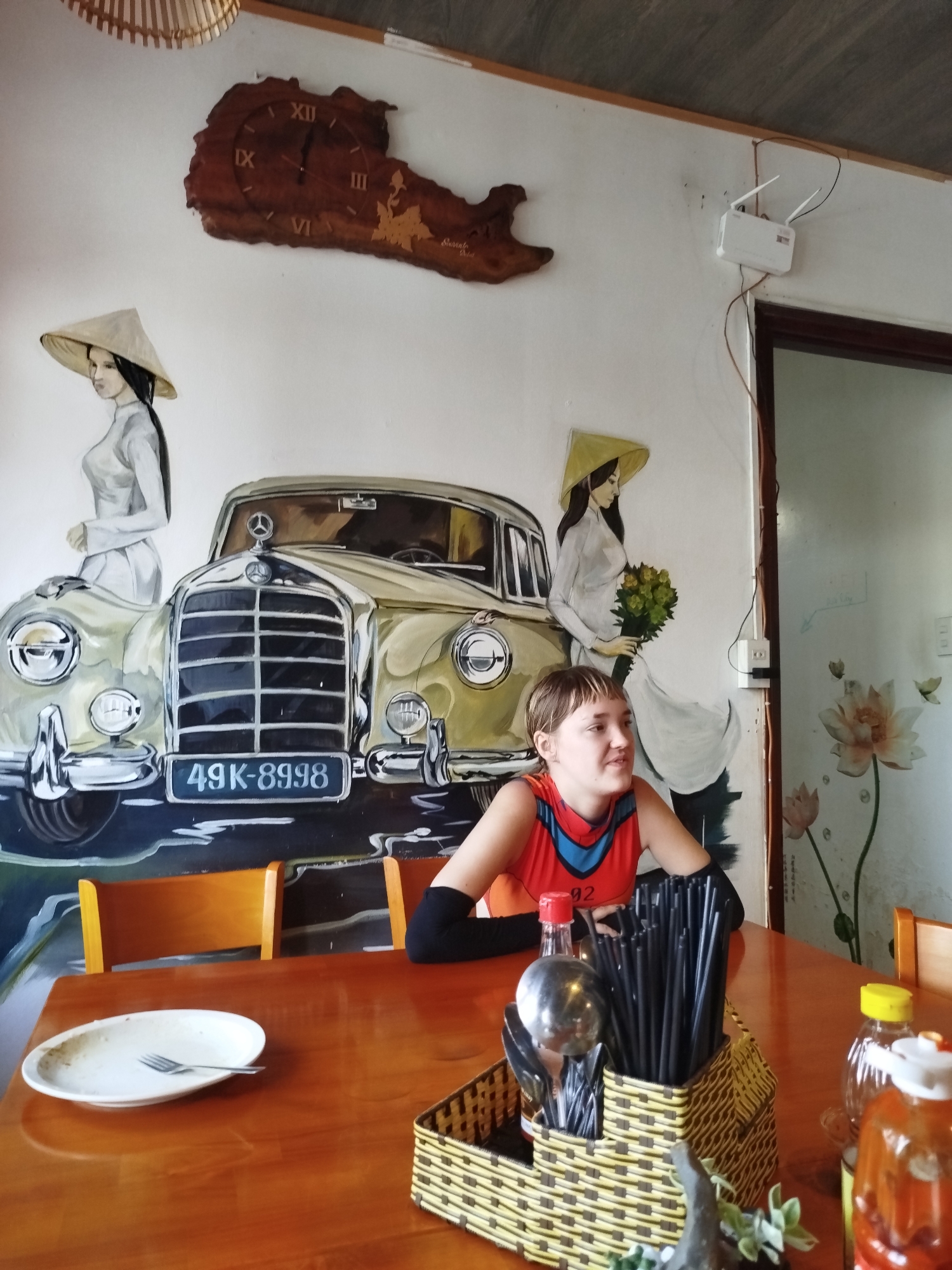 A slightly high-angle indoor shot captures a person with short hair seated at a wooden table in what appears to be a restaurant. The person, wearing a red and blue top and black sleeves, looks slightly to the right. On the table, there is a plate with a fork, several bottles of condiments, and a basket containing chopsticks and cutlery.
The wall behind the person features a mural. The mural depicts two women in traditional Vietnamese conical hats and white Áo dài dresses flanking a classic Mercedes-Benz car. The car's license plate reads "49K-8998." Above the mural is a wooden clock shaped like a geographical feature. To the right of the mural and near the top of the image, there's a WiFi router mounted on the wall.
The scene strongly suggests the location is in Da Lat, Vietnam, based on the cultural elements such as the traditional clothing and the mural depicting the city's common vintage car motifs. The environment is typical of a local restaurant, with simple wooden furniture and decorative elements reflecting the region's identity.
