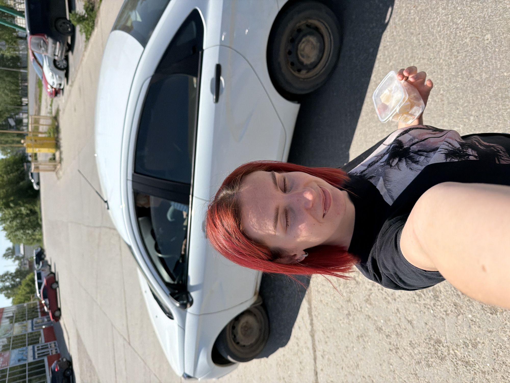 A woman with red hair is taking a selfie in front of a white car in Chaikovsky, Russia. She is holding a small, clear plastic container in her hand, which appears to contain small, white food items. Other vehicles are parked nearby, and there is a building in the background with visible advertising. The ground is paved.