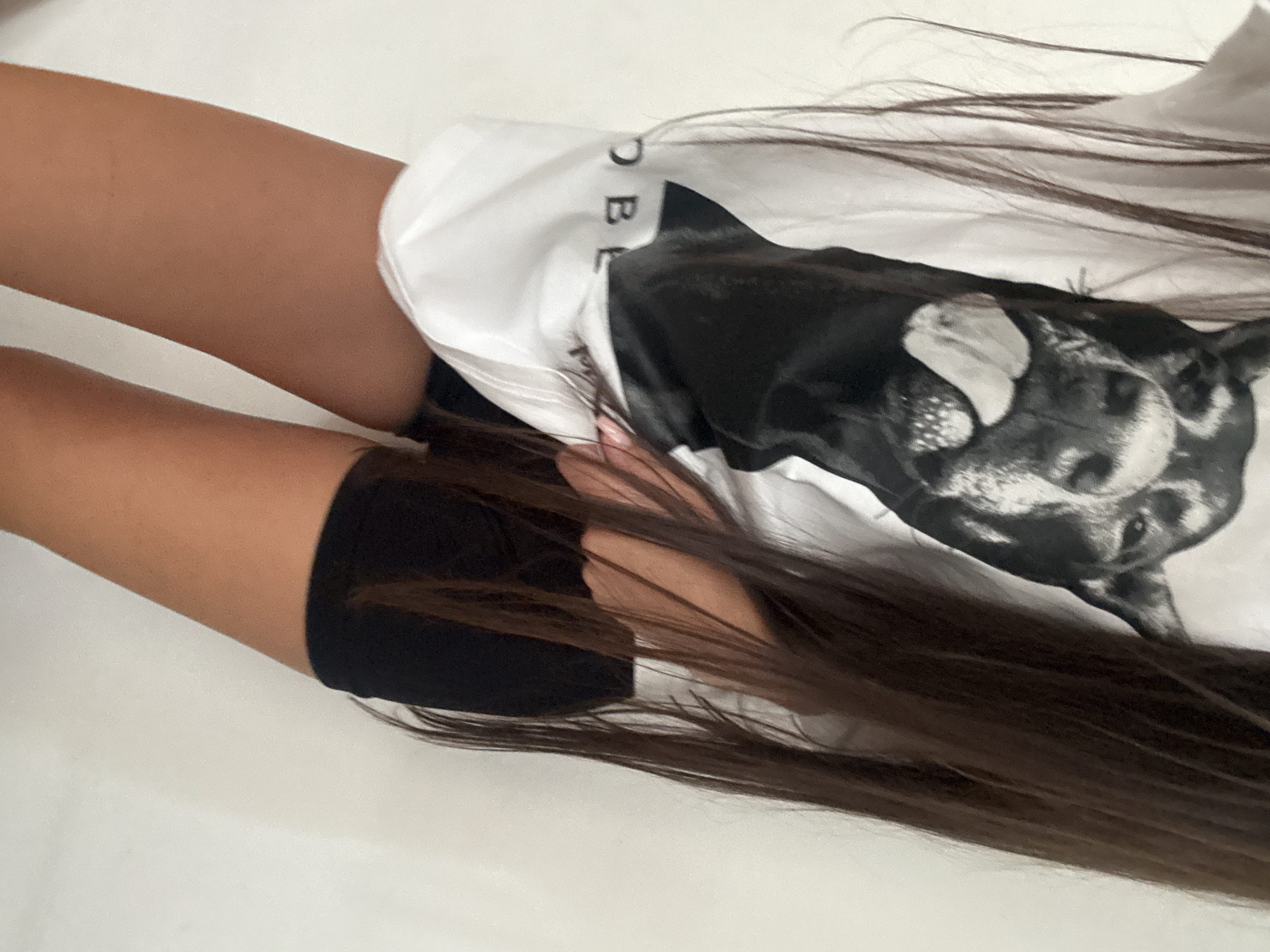 The image shows a young woman standing against a plain white wall. She has long, dark brown hair that falls down her back and partially obscures her face and shoulders. She is wearing an oversized white t-shirt with a black and white graphic of a dog's face on the front. The dog appears to be a Doberman Pinscher with its tongue sticking out. The t-shirt is partially tucked into a pair of black, high-waisted shorts. Her legs are tanned, and she appears to be indoors in a well-lit environment. There are no discernible elements in the image that suggest a specific location within the city of Chișinău, Moldova, or any other city. The background is a featureless white wall.
