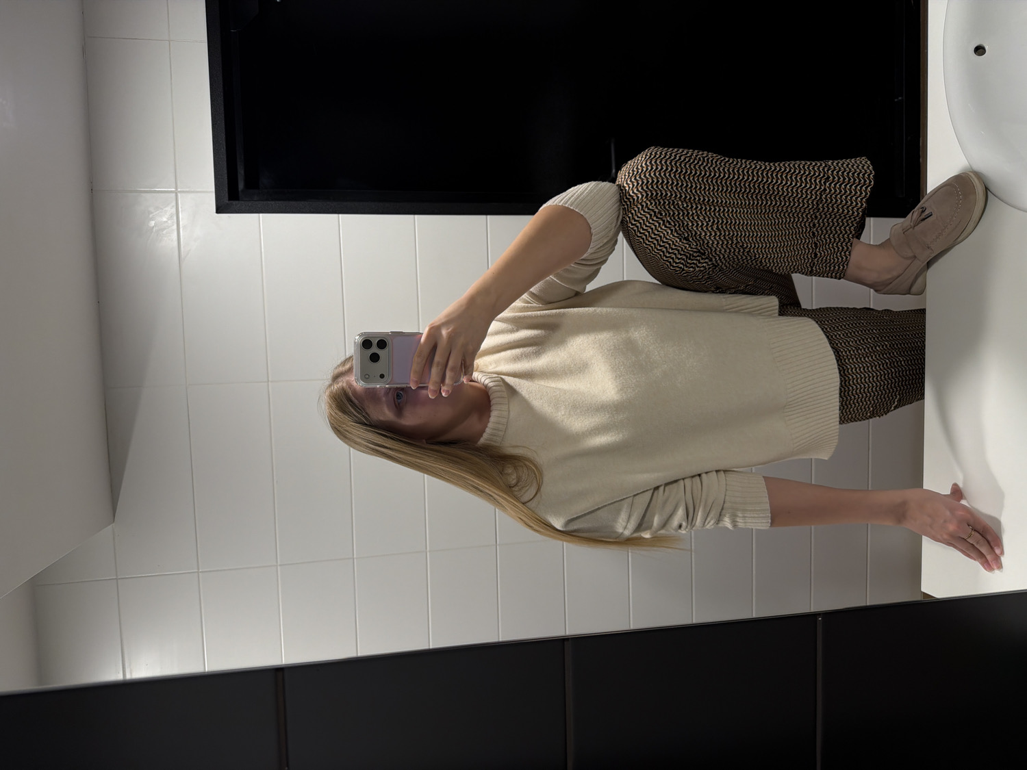 A mirror selfie captures a person from approximately the mid-thigh up, standing in a brightly lit modern bathroom in Izhevsk, Russia. The person, likely female, has long, straight blonde hair and is looking towards the screen of a light purple smartphone, which she holds with her right hand to take the photo. The phone features a prominent triple-lens camera module.
She is dressed in a comfortable, light beige or cream knit sweater with ribbed cuffs and a matching ribbed hem. Her pants feature a distinctive patterned design, appearing as a zigzag or chevron motif in alternating dark and light brown colors. On her feet are light brown, suede-like loafers with a tassel detail, resting near a white ceramic surface, likely a sink.
The background consists of white square tiles on the wall, separated by dark grout lines, creating a clean and geometric aesthetic. Above the person's reflection, a dark, rectangular fixture or frame is visible, and a similar dark, horizontal surface runs along the bottom of the mirror. The overall setting suggests a public or commercial restroom. The scene is well-lit with ambient light, and there is no visible text. The person's expression is neutral, focused on taking the selfie. No specific time of day or weather is discernible.