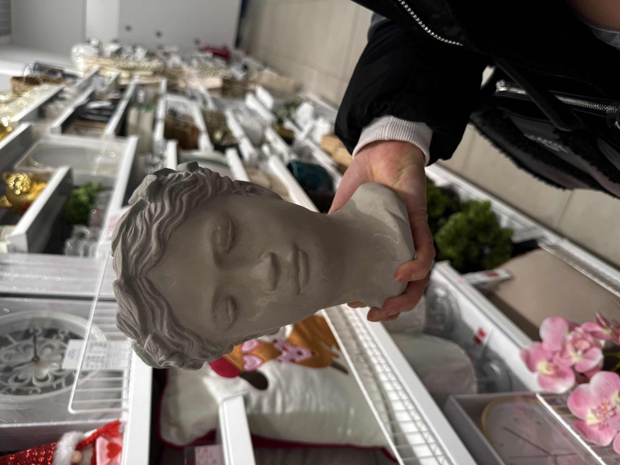 A person holds a gray concrete bust of a head in their hand while standing in a store with shelves stocked with various items. The person is wearing a black jacket with a white sweater underneath. The shelves contain decorative items such as clocks, flowers, greenery, and other ornaments. The location could possibly be in the city of Stara Zagora, Bulgaria.