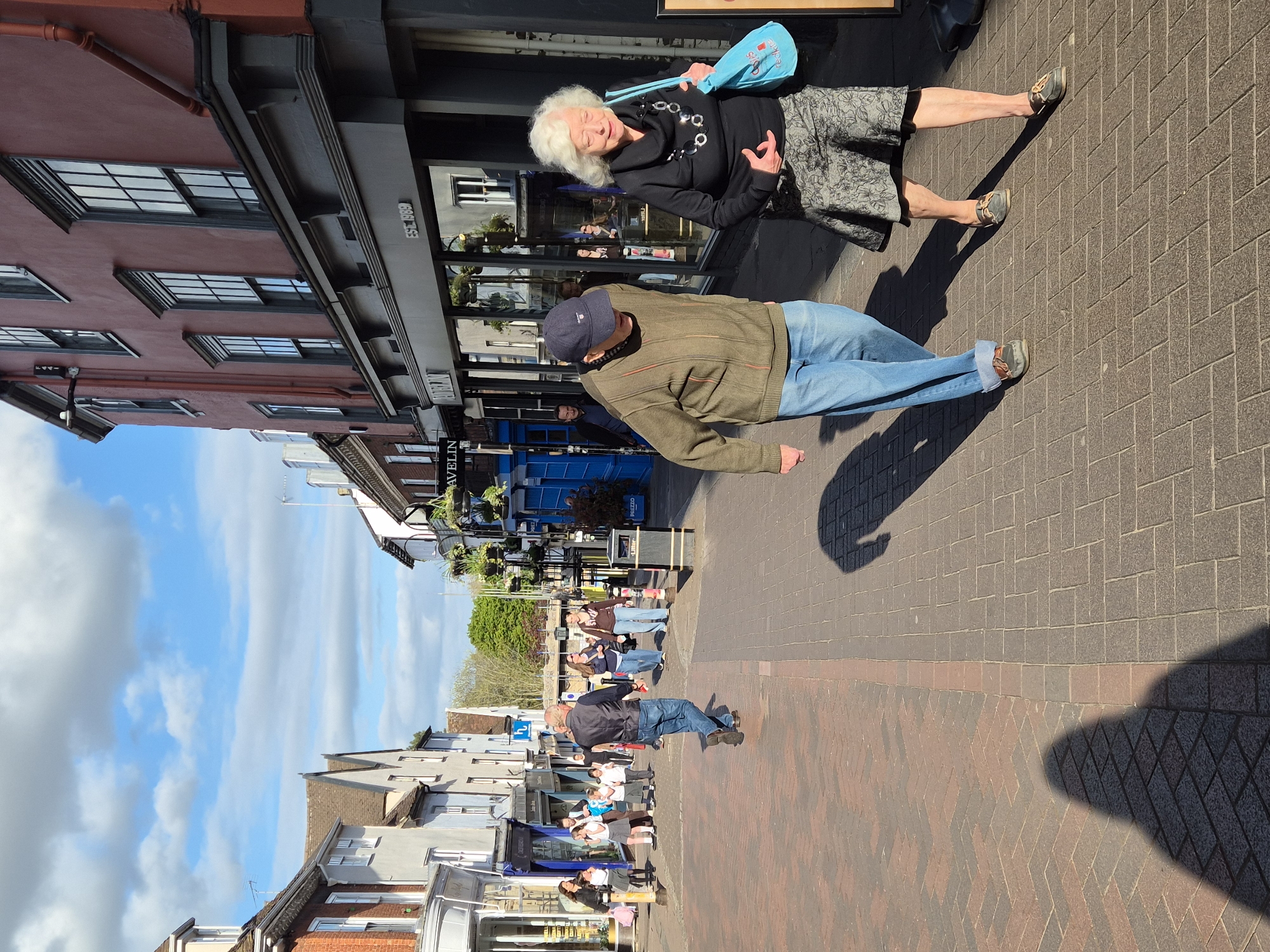 The image shows a street scene in West Suffolk, United Kingdom, likely a pedestrianized area. Two people, an elderly woman with white hair and a younger man in a blue baseball cap, stand on a brick-paved street in front of a building. The woman wears a black top and patterned skirt, while the man wears a green jacket and blue jeans. Further down the street, other people are walking. Buildings line both sides of the street, with shops and businesses visible. The sky is blue with scattered clouds.