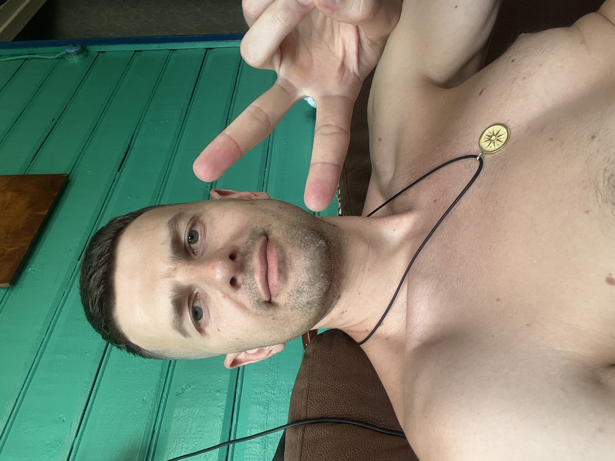 A man with short brown hair and stubble is pictured from the chest up, seated in front of a green wooden wall. He is shirtless and wears a black cord necklace with a gold pendant. His right hand is raised, fingers forming a peace sign. The man appears to be indoors, with a brown upholstered object visible to his left. The green wall is composed of horizontal planks, and a wooden panel is mounted above and to the left of the man's head. The scene is lit by natural light. This image was captured in Navahrudak, Belarus.