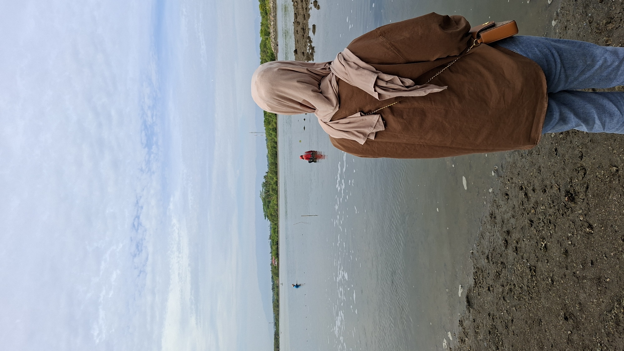 The image depicts a person wearing a brown shirt and a light brown hijab standing with their back to the camera, looking out over a body of water. In the distance, two individuals can be seen in the water. One is wearing a blue hat and the other is wearing a red shirt. The background features a treeline and hills under a cloudy sky. The foreground consists of a muddy shoreline with some debris. The scene appears to be a coastal area or a large body of water, possibly near Lhokseumawe, Indonesia, given the context. The overall atmosphere is overcast and calm.