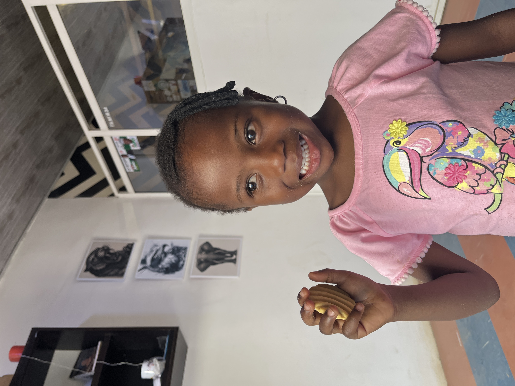 A young girl stands indoors, holding a cookie. She is smiling, wearing a pink shirt with a bird design. There is a black shelf, three pictures on a wall, and a window. The location is Jos, Nigeria.