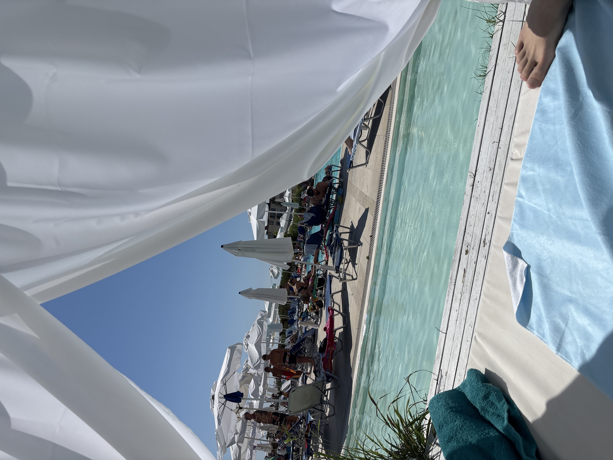 The image captures a sun-drenched outdoor scene at a resort or hotel in Byala, Bulgaria. In the foreground, a white canopy drapes across the top of the frame, partially obscuring a clear blue sky. Below the canopy, a person's foot is visible resting on a light-colored lounge pad, with a blue towel and a teal towel placed nearby. The poolside area is laid with light wooden planks.
Beyond the immediate foreground, a vibrant turquoise swimming pool stretches across the midground. Along the pool's edge, rows of empty and occupied lounge chairs are neatly arranged under numerous white umbrellas, providing shade for sunbathers. Several individuals are seen relaxing on these chairs or walking around the pool area. In the background, a few buildings with balconies are visible, suggesting the presence of accommodations. The overall atmosphere is one of leisure and relaxation, typical of a summer vacation destination.