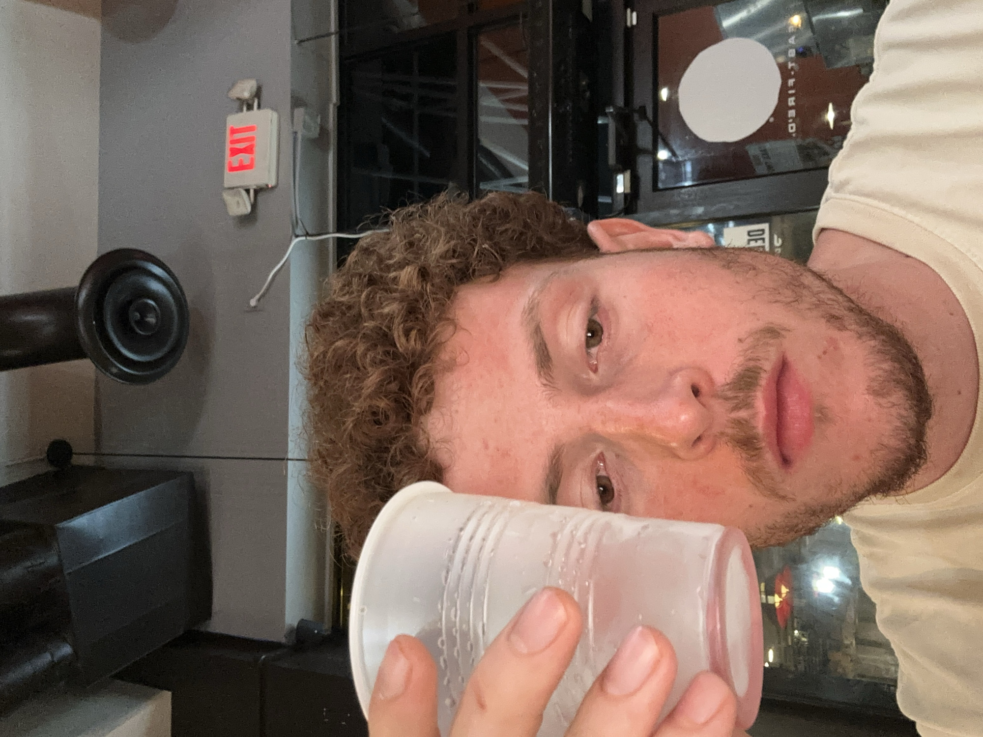 The media file displays an image rotated 90 degrees counter-clockwise. The primary subject is a man with curly brown hair and a beard, whose face is seen in profile, looking towards the top of the frame. He holds a clear plastic cup, showing condensation and water droplets, to his mouth, appearing to drink from it. He is wearing a light-colored t-shirt.
In the background, on a grey wall to the left of the man's head, a white "EXIT" sign with red illuminated text is visible, accompanied by two small emergency lights. Below the sign, a black, old-fashioned telephone receiver or speaker is mounted on the wall. To the lower left, a dark, box-like object is partially visible.
Above and behind the man's head, a window reflects a large white circle and some reversed text, which appears to include "D.R-T.". Dark structural elements are also reflected in the window. The setting suggests an indoor public or commercial space, possibly a bar or restaurant, given the "EXIT" sign and interior details. The man's expression is neutral. No specific time of day or weather is discernible.