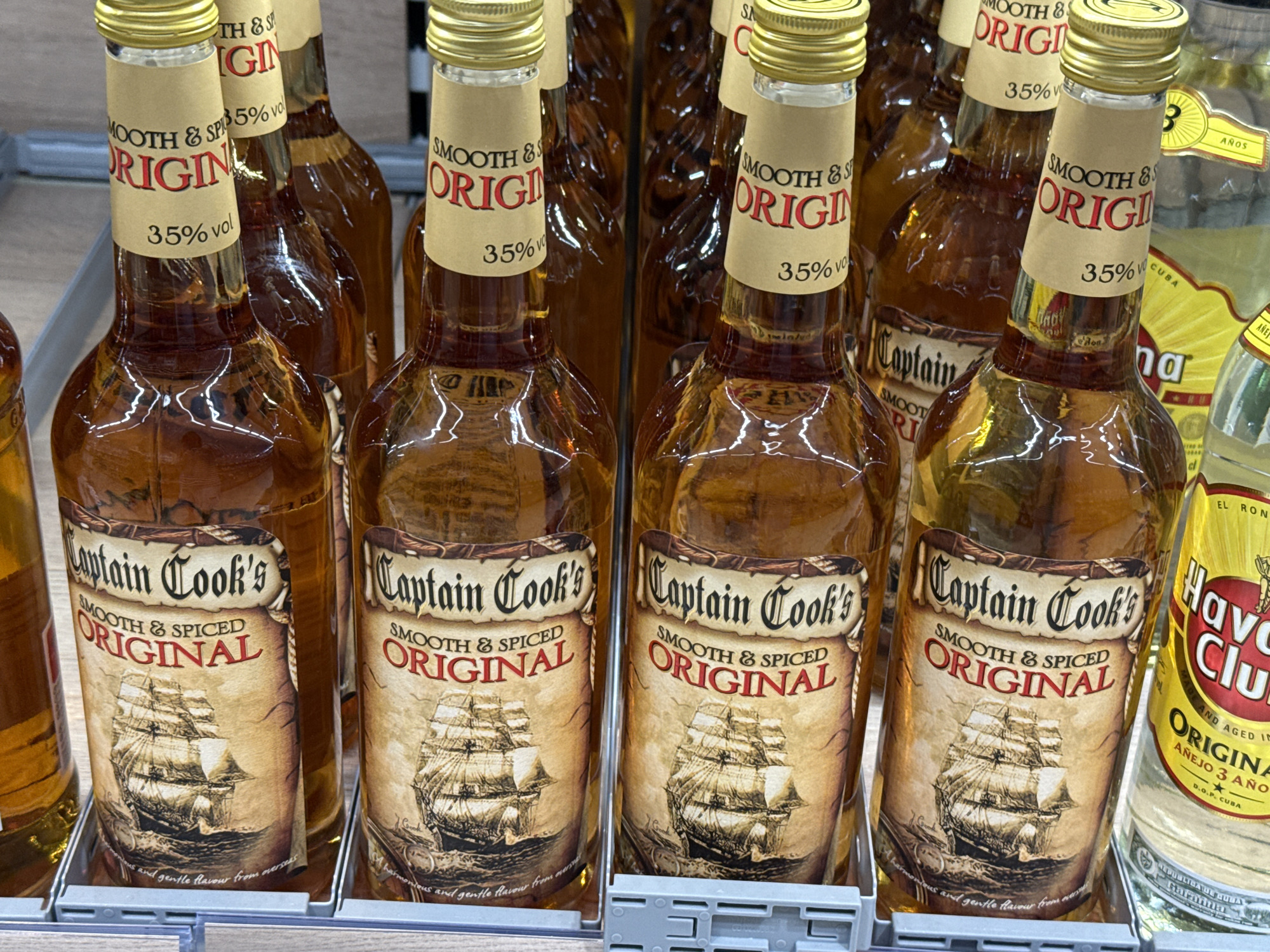 The image shows a close-up, eye-level view of several bottles of Captain Cook's Smooth & Spiced Original liqueur on a shelf. The bottles are made of clear glass and filled with amber-colored liquid. Each bottle has a distinctive label that features a ship illustration and the text "Captain Cook's SMOOTH & SPICED ORIGINAL" along with "35% vol". The labels are designed to look like aged parchment. The bottles are arranged in rows, with the focus on the front row where four bottles are clearly visible. Behind these, more bottles can be seen. To the right of the Captain Cook's bottles, partially visible, are bottles of Havana Club rum, distinguishable by their yellow and red labels. The shelf is a standard retail display shelf, likely in a supermarket or liquor store. The lighting is artificial and appears to be from above, creating reflections on the glass bottles. The background is blurred, suggesting it is the interior of a commercial establishment. There are no people or animals visible in the image. The overall impression is a product display in a retail setting. This image is from a store in Herzberg, Germany.