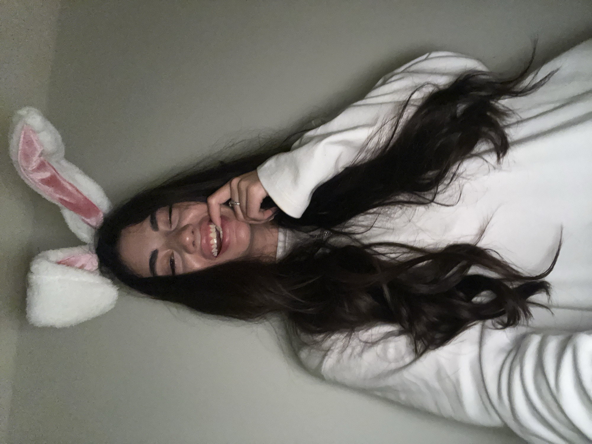 A young woman with long, dark hair is wearing white bunny ears and a white sweatshirt. She is smiling and has her finger near her mouth. The background is a plain, light-colored wall. No information about the location in Chotěšov, Czechia is provided in the image.
