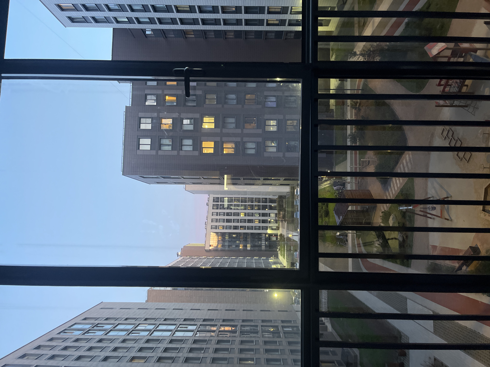 The image captures a view from inside a building, looking out through a window onto a modern residential complex during twilight. A dark window frame, featuring vertical and horizontal mullions, dominates the foreground, with a window handle visible on the left side (when viewed upright).
Beyond the window, multiple high-rise apartment buildings with contemporary architecture are visible. These buildings have uniform grey or dark grey facades and numerous rectangular windows. Many of the windows are illuminated with warm yellow light, strongly suggesting the time of day is evening or night. The sky above is a dim, light blue-grey, consistent with dusk.
Below the buildings, a well-maintained courtyard or shared community space is visible. This area features paved walkways, some with a reddish-brown color, contrasting with lighter paths and green landscaped sections. A children's playground is clearly visible through the window bars on the right, including structures with a red roof, metal climbing frames, and other ground-level play equipment.
No people are visible in the scene. The overall impression is a quiet, urban residential environment, characteristic of modern developments in a city like Saint Petersburg, Russia. There is no visible text in the image.