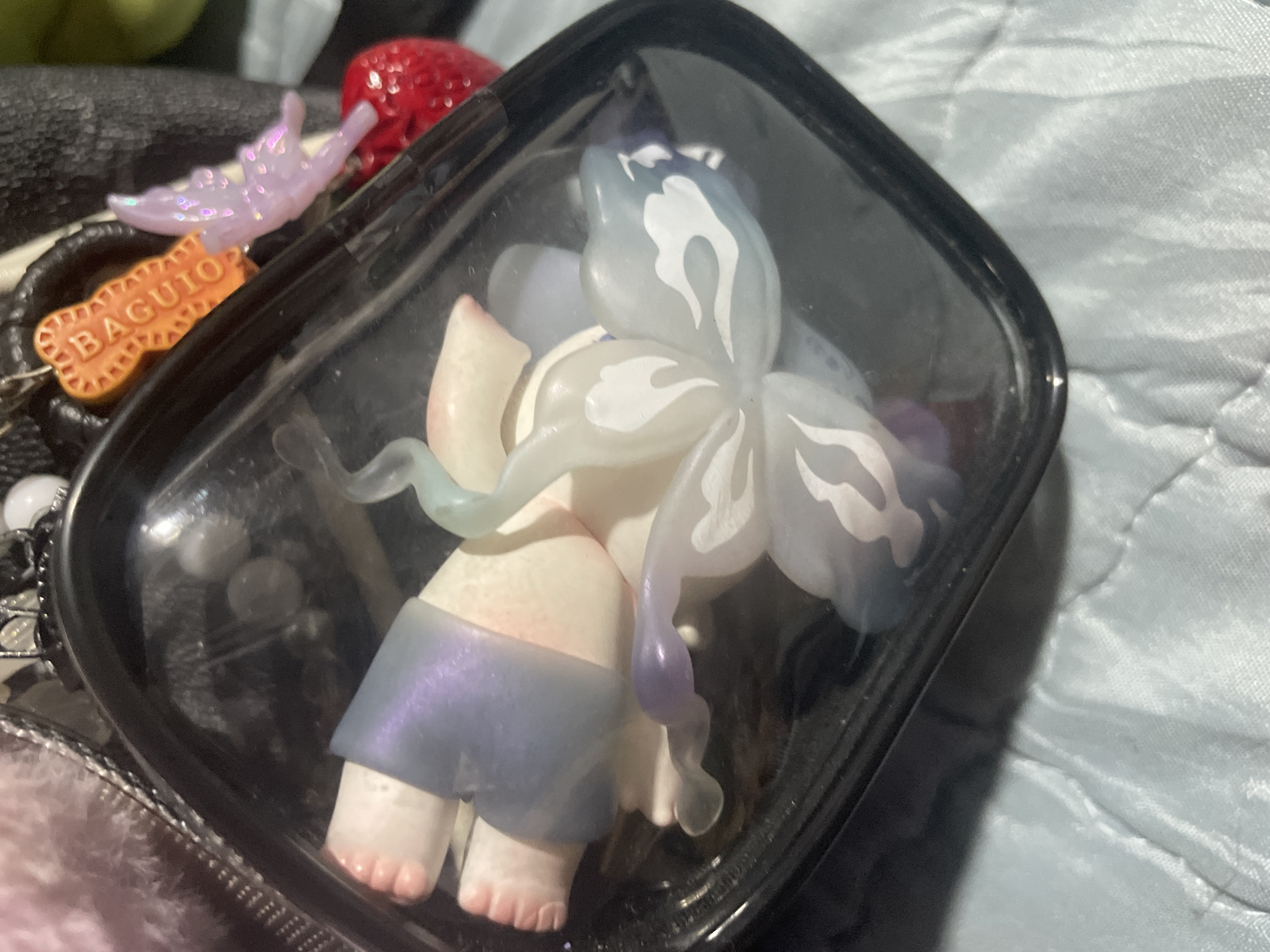 The image, captured in Davao City, Philippines, displays a close-up view of various decorative items. A black-rimmed, clear plastic container with rounded corners occupies the central frame. Inside, two primary subjects are visible: a small, light-skinned figurine and a multi-petaled floral object. The figurine, positioned on its back, features prominent, articulated feet and legs covered by translucent, shimmering purple material. Its torso is pale and smooth, with the head mostly out of frame to the left. Adjacent to the figurine is a stylized floral object composed of translucent white, light blue, and light purple petals. Several petals display intricate white patterns resembling swirling clouds or flames.
Surrounding the container, elements of the immediate environment are visible. In the upper left, a soft, light pink, textured material, possibly faux fur, is present. At the upper right, a collection of charms is suspended. These include an orange, rectangular, biscuit-shaped charm with the word "BAGUIO" embossed in white text and serrated edges. Next to it hangs a translucent, iridescent purple butterfly charm, which is attached to a shiny, textured red sphere resembling a cherry. The container and charms rest on a light blue, quilted textile surface, likely a blanket or sheet, which fills the lower portion of the frame. The scene is well-lit, with reflections visible on the clear plastic surface of the container.