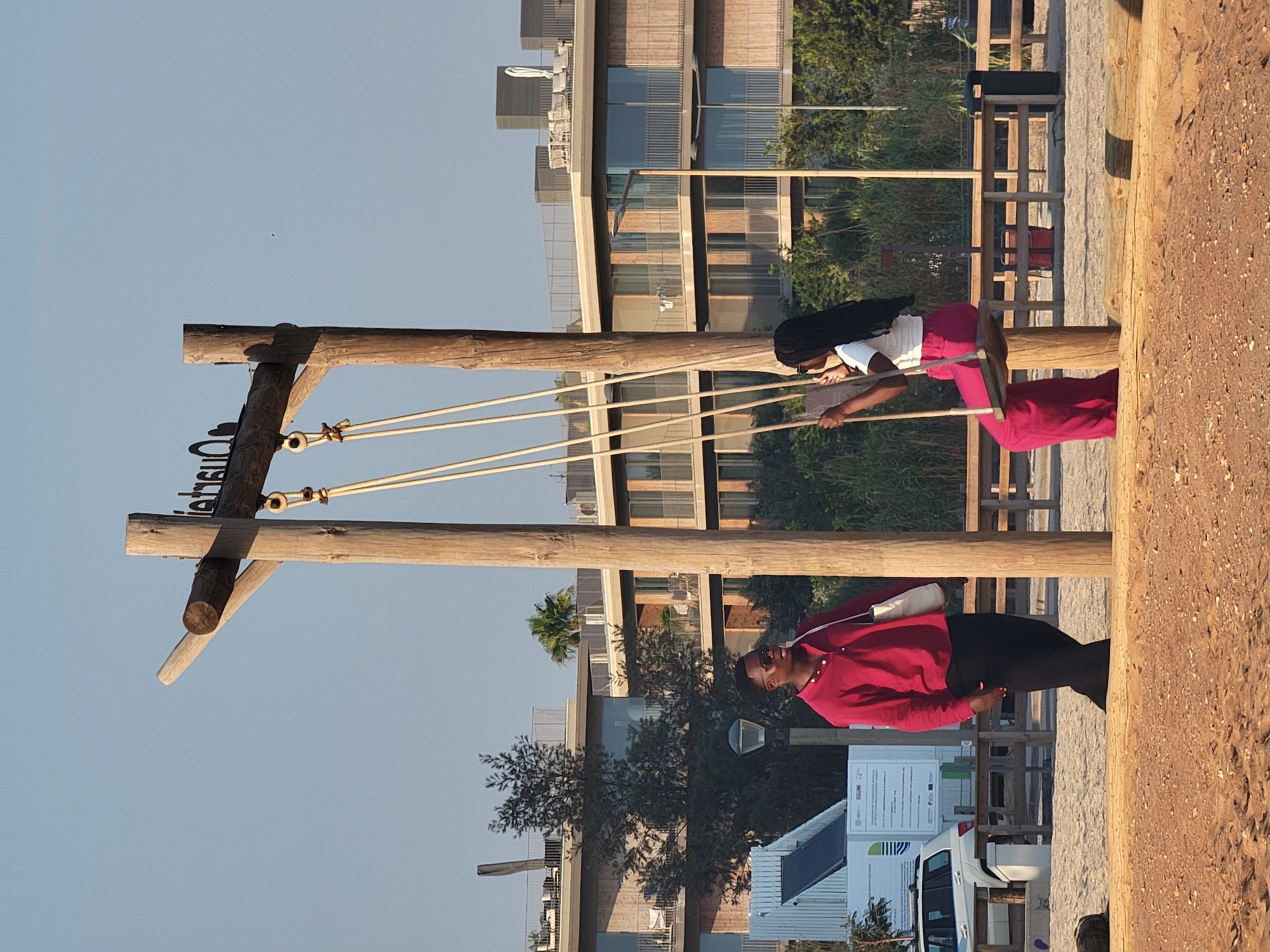 The image depicts a beach scene in Quarteira, Portugal, with a wooden swing set as a central element. On the left, a Black man wearing a red shirt and black pants stands looking to the right, a white bag slung over his shoulder. To the right, a Black woman with braided hair is seated on the swing, wearing a white top and bright pink wide-leg pants, looking to her left and holding a book or phone. Behind them, a modern, multi-story building with a light-colored facade and balconies is visible, surrounded by trees and vegetation. A white car and a kiosk with solar panels are also present in the background, on the sandy beach. The sky is clear and blue, suggesting fair weather.
