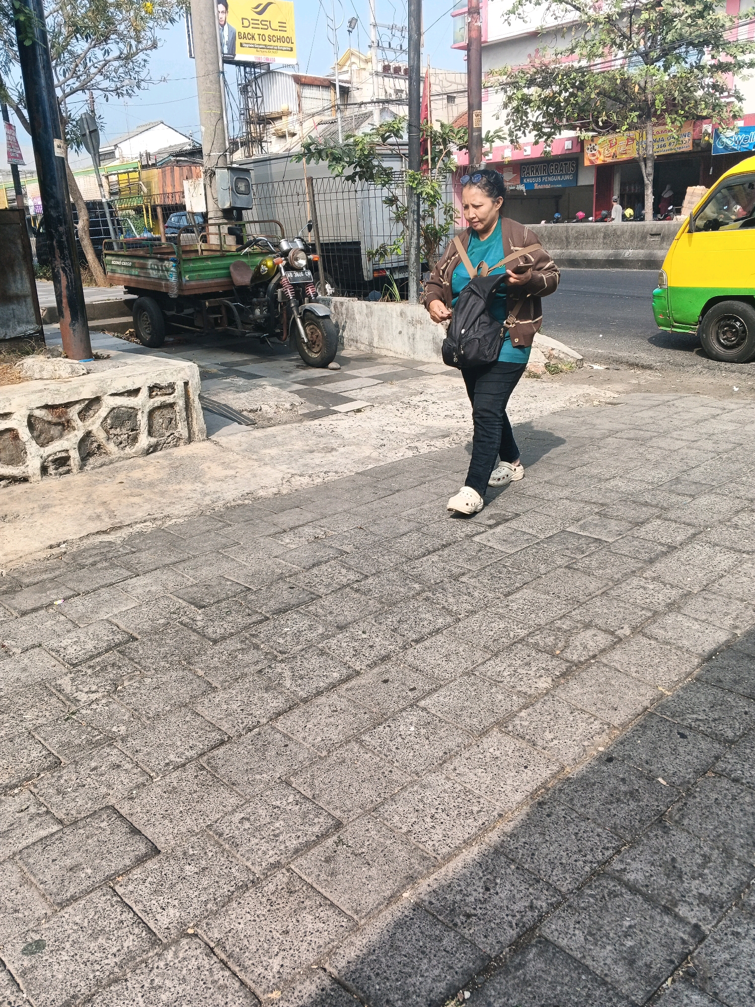 A woman is walking on a cobblestone sidewalk in Ungaran, Indonesia. She is wearing a brown jacket over a teal shirt, black pants, and white clogs. She has a black backpack on her back and is holding her phone in her left hand. The sidewalk is bordered by a low stone wall on the left. In the background, there is a green and yellow vehicle, a motorcycle with a cart attached, and several buildings. The sky is clear and the sun is shining.