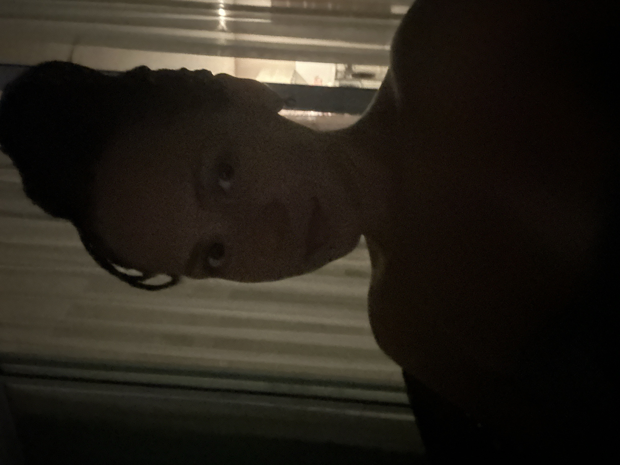 A person with braided hair in a bun is facing the camera. The person is partially visible in a dark setting with what appears to be window blinds in the background. The location cannot be determined to be Pattaya City, Thailand based on the provided image alone.