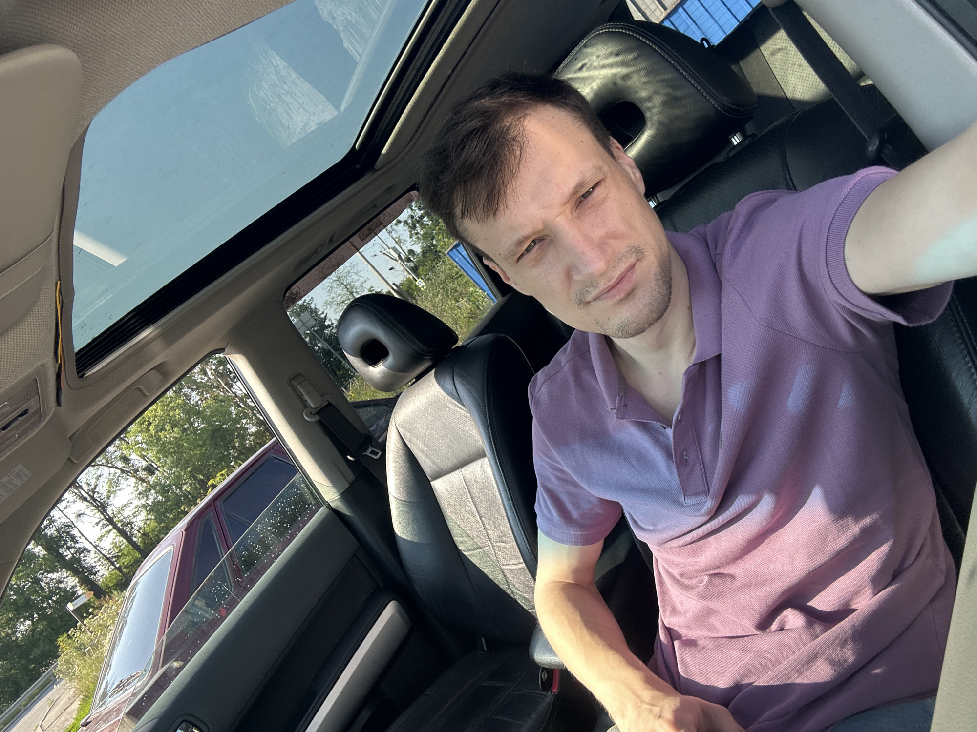 A man is taking a selfie inside a car. He is wearing a purple polo shirt and has short brown hair and a stubble beard. The car has a sunroof that is open, revealing a clear blue sky. The interior of the car appears to be black leather. Outside the car, trees and another car can be seen. The location is the city of Shatava, Ukraine.