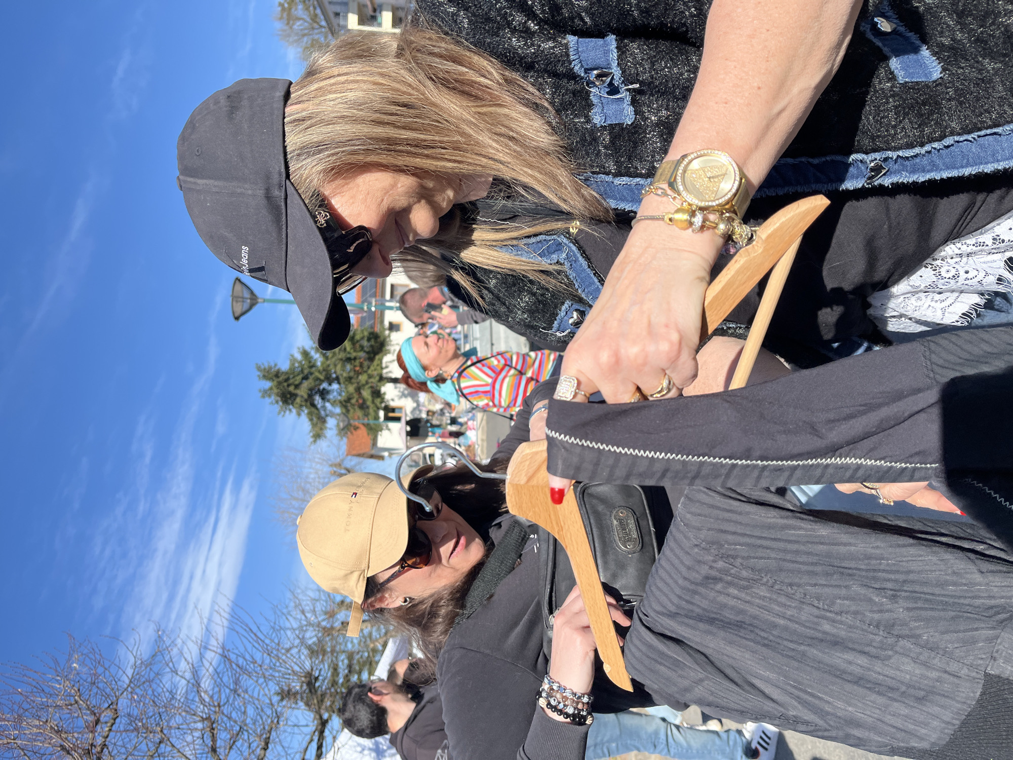 Two women examine clothing at an outdoor market on a sunny day in Bellegarde-en-Forez, France. One woman, wearing a black jacket with denim trim, a gold watch, and a black baseball cap, holds a wooden hanger with a piece of clothing. The other woman, wearing a tan baseball cap and sunglasses, also holds a garment on a wooden hanger. Other people are visible in the background. The scene is outdoors with a blue sky and scattered clouds.