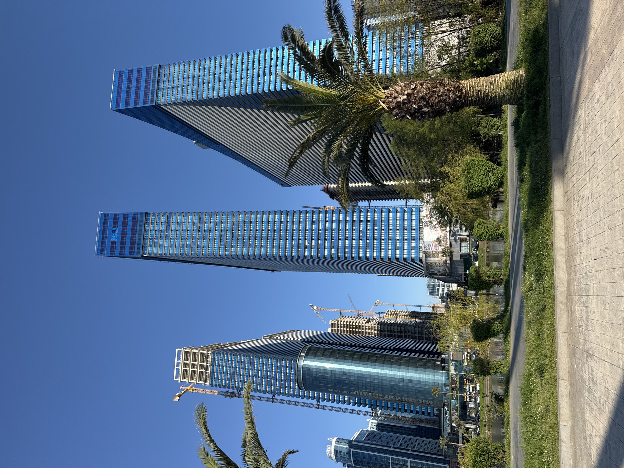 This image captures a view in Batumi, Georgia, looking upward at several modern skyscrapers under a clear blue sky. The architecture is characterized by blue glass facades with distinctive horizontal lines. Construction cranes are visible, indicating ongoing development in the area. A palm tree stands nearby, juxtaposing the natural element with the urban landscape. A sidewalk and strip of greenery are visible in the foreground.