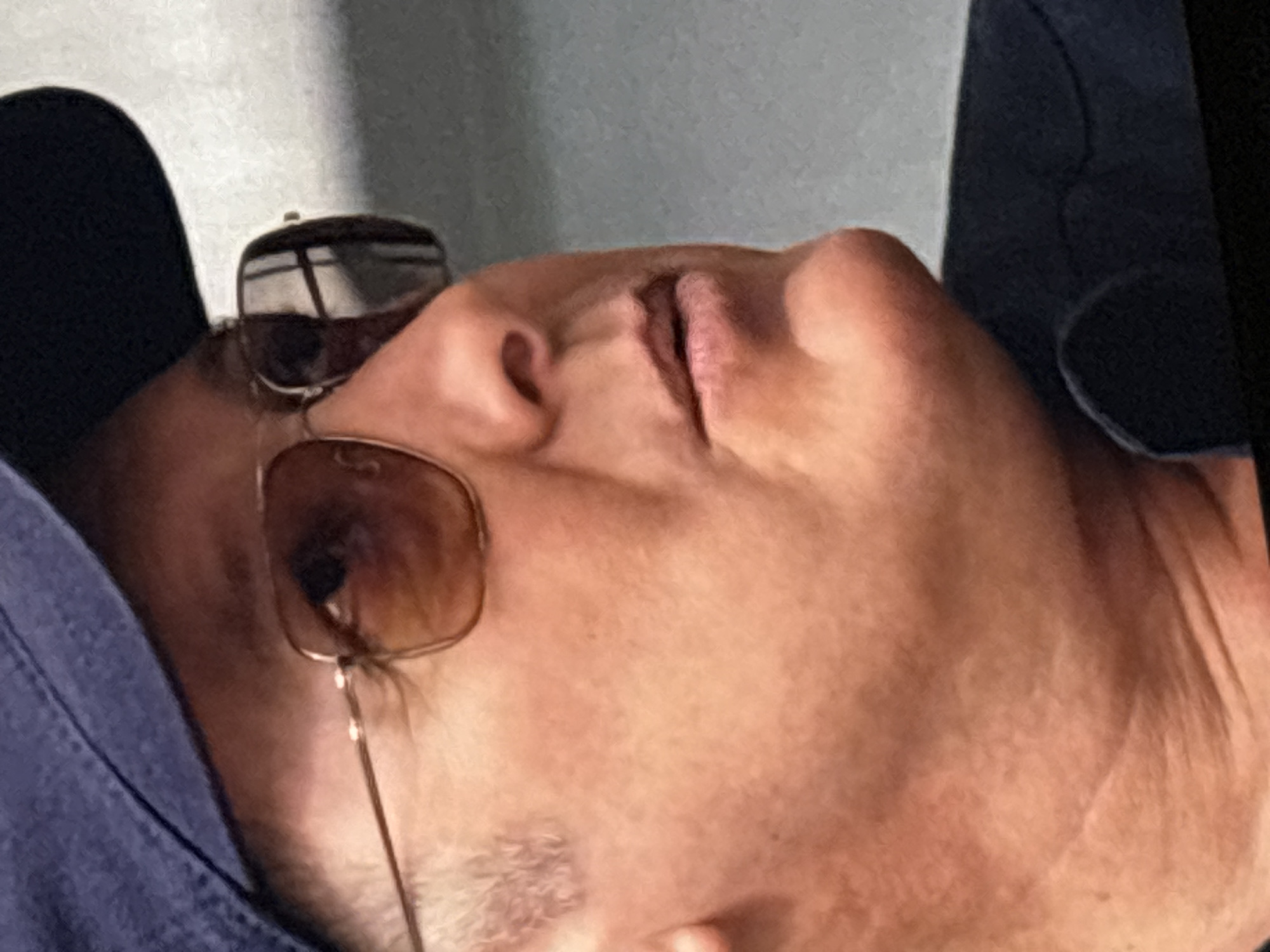 This close-up, rotated image, set in Budva, Montenegro, captures a person, likely male, in a state of rest or sleep. The individual's head is viewed from a side profile, with their face angled upwards, revealing their nose, mouth, and chin. A pair of aviator-style sunglasses are perched on their face, obscuring their eyes. The person's mouth is slightly agape, suggesting deep relaxation or sleep. Light stubble is visible on their cheek and jawline. A blue garment, possibly a shirt or jacket, is draped over their shoulder and chest. The background is softly blurred, showing indistinct light-colored surfaces, indicative of an indoor or shaded outdoor setting with diffused lighting. The scene conveys a peaceful and serene atmosphere. No discernible text is present in the image.