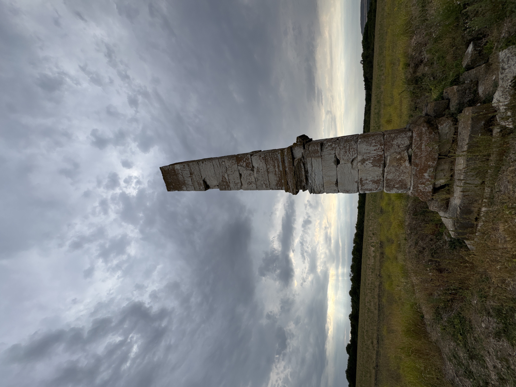 In the city of Lesicheri, Bulgaria, an ancient, weathered stone obelisk stands in an open field under a dramatic, cloudy sky. The obelisk is constructed from large, stacked stone blocks, showing signs of erosion and moss growth, giving it an aged appearance. It is tilted slightly, leaning to the right. The surrounding environment consists of a grassy field with patches of dry vegetation. In the background, a line of trees forms a dark silhouette against the horizon, beyond which hills are faintly visible. Sunbeams pierce through the clouds in the distance, creating shafts of light that illuminate the field. The scene is captured from a low angle, emphasizing the height and imposing nature of the obelisk against the vast sky.