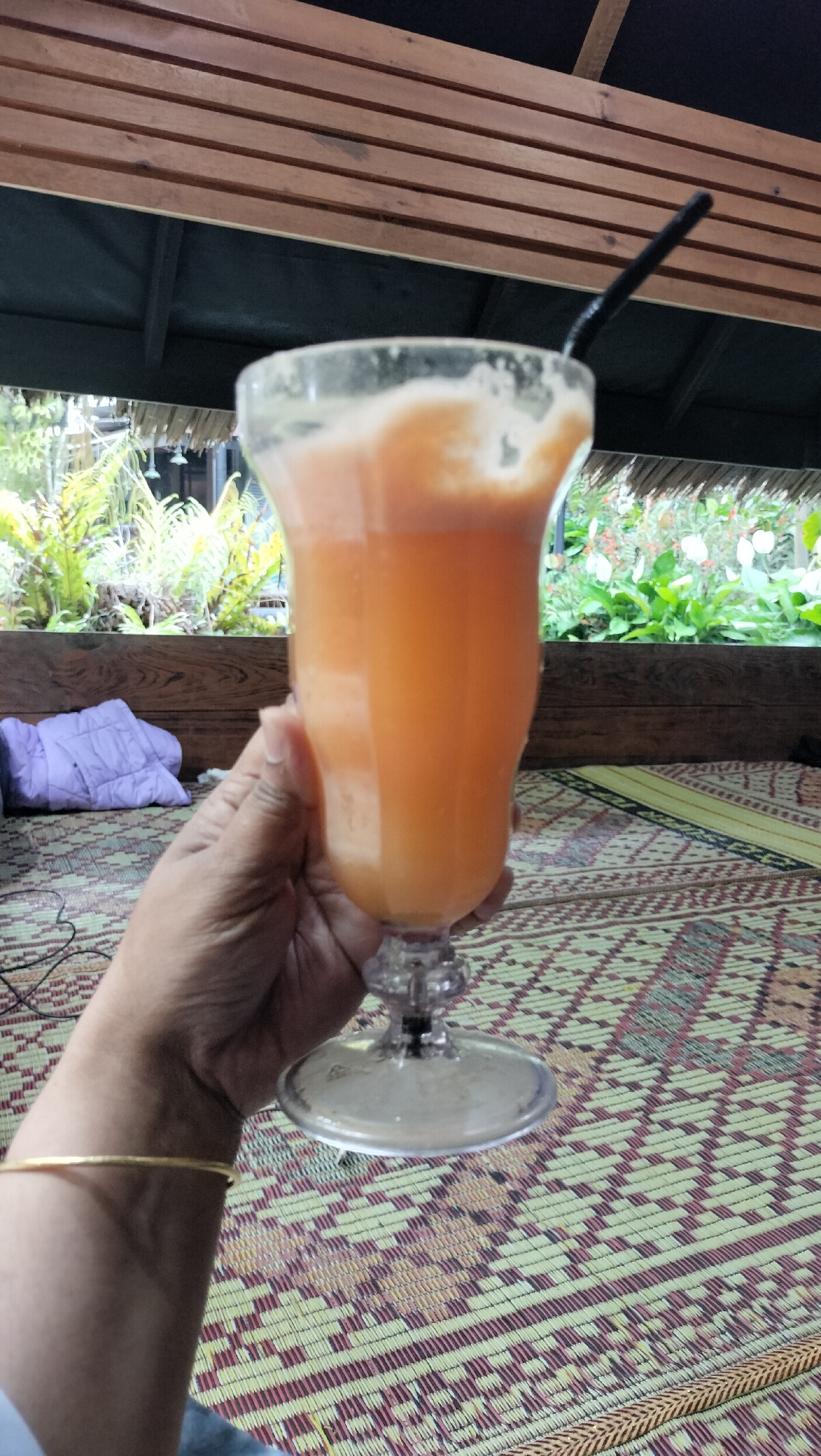 A hand is holding a clear glass filled with an orange beverage and a black straw. The glass has a stem and a wide base. The hand wears a thin gold bracelet on the wrist. The background shows lush green foliage and a wooden structure with parallel wooden slats overhead. A patterned woven mat covers the surface where the hand and glass are positioned. The scene appears to be outdoors, possibly in a cafe or restaurant, with a view of a garden or natural landscape. The image does not provide any specific information to determine a location in the city of Tansaril, Indonesia.