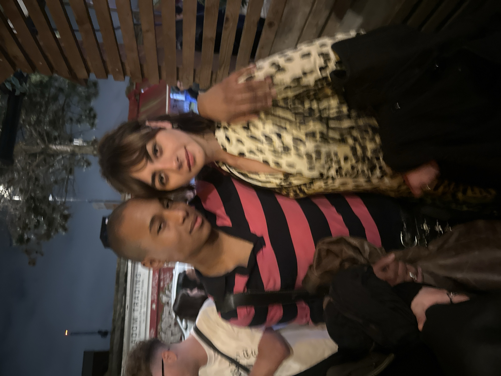 The image shows two people posing closely together. One person has a shaved head, a pink and black striped shirt, and is positioned lower in the frame. The other person has shoulder-length brown hair with bangs and is wearing a patterned shirt. Other people are present in the background, some blurred out. The location appears to be outdoors, with a building in the background that may read "LONDON TRADING." Wooden beams are visible overhead. The image is taken at night. The precise location in London cannot be determined with certainty, but the sign suggests an area associated with trading or commercial activities.