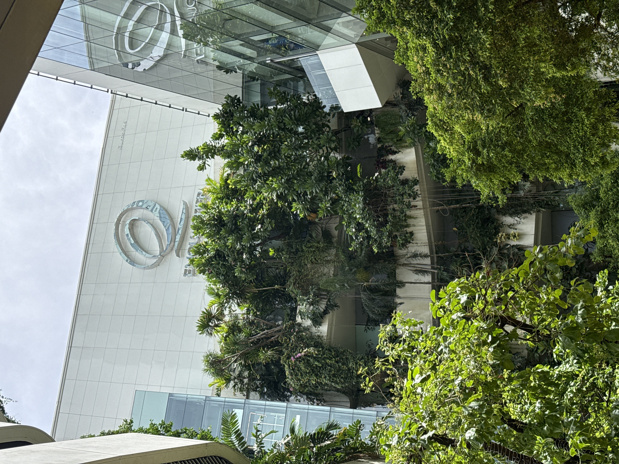 The image shows a view looking up at the Siam Paragon shopping mall in Bangkok, Thailand. A large silver "@" logo is visible on the white facade of the building. Lush greenery, including trees and shrubs, partially obscures the view of the building's exterior, suggesting a garden or landscaping feature. The presence of reflective glass and a modern architectural style are also evident.