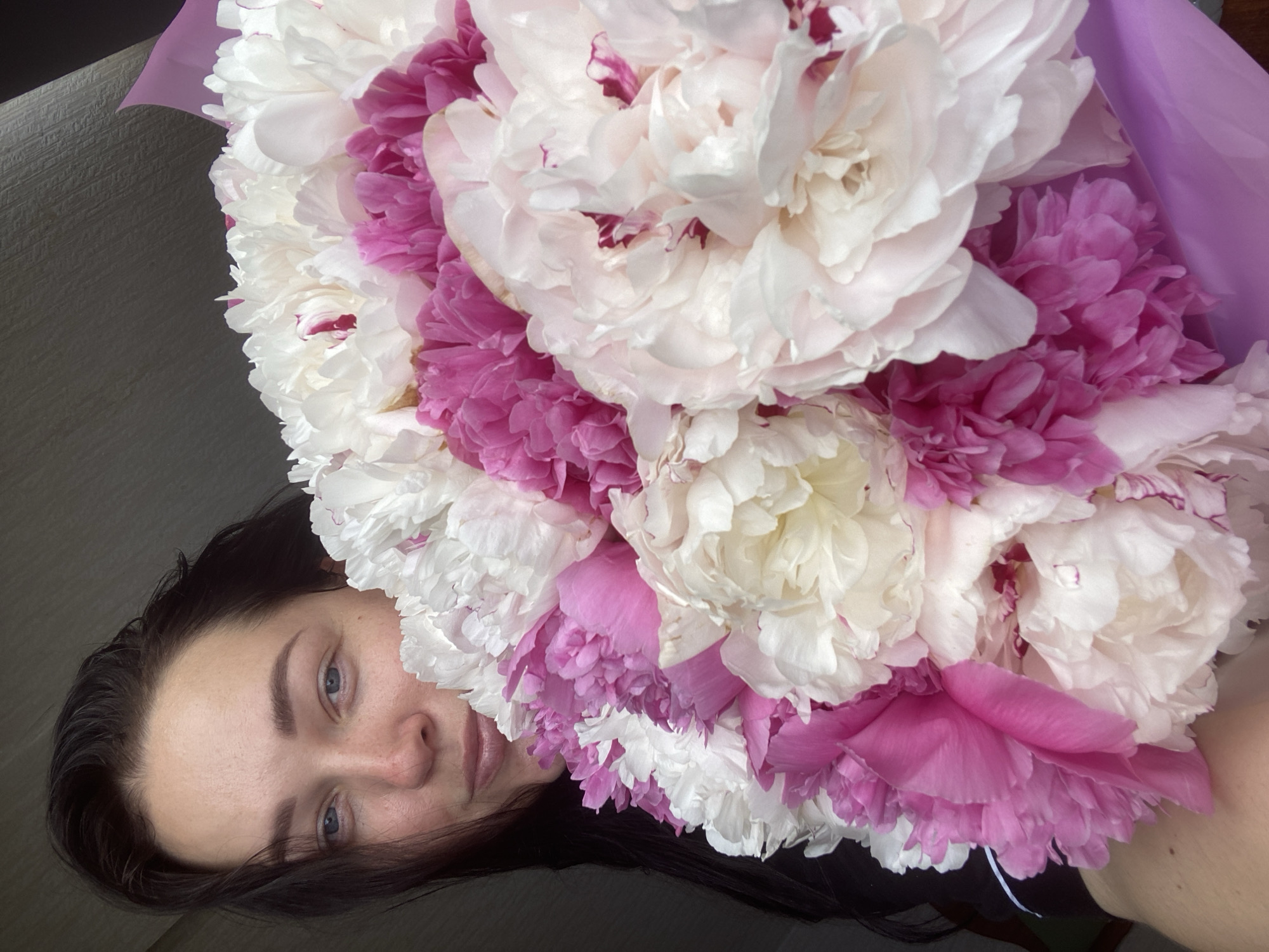 A woman with dark hair and blue eyes is lying down with a bouquet of white and pink peonies. The flowers are clustered together and wrapped in light purple paper. The woman's face is partially obscured by the flowers. The background is a blurred, neutral color. The location is unknown.
