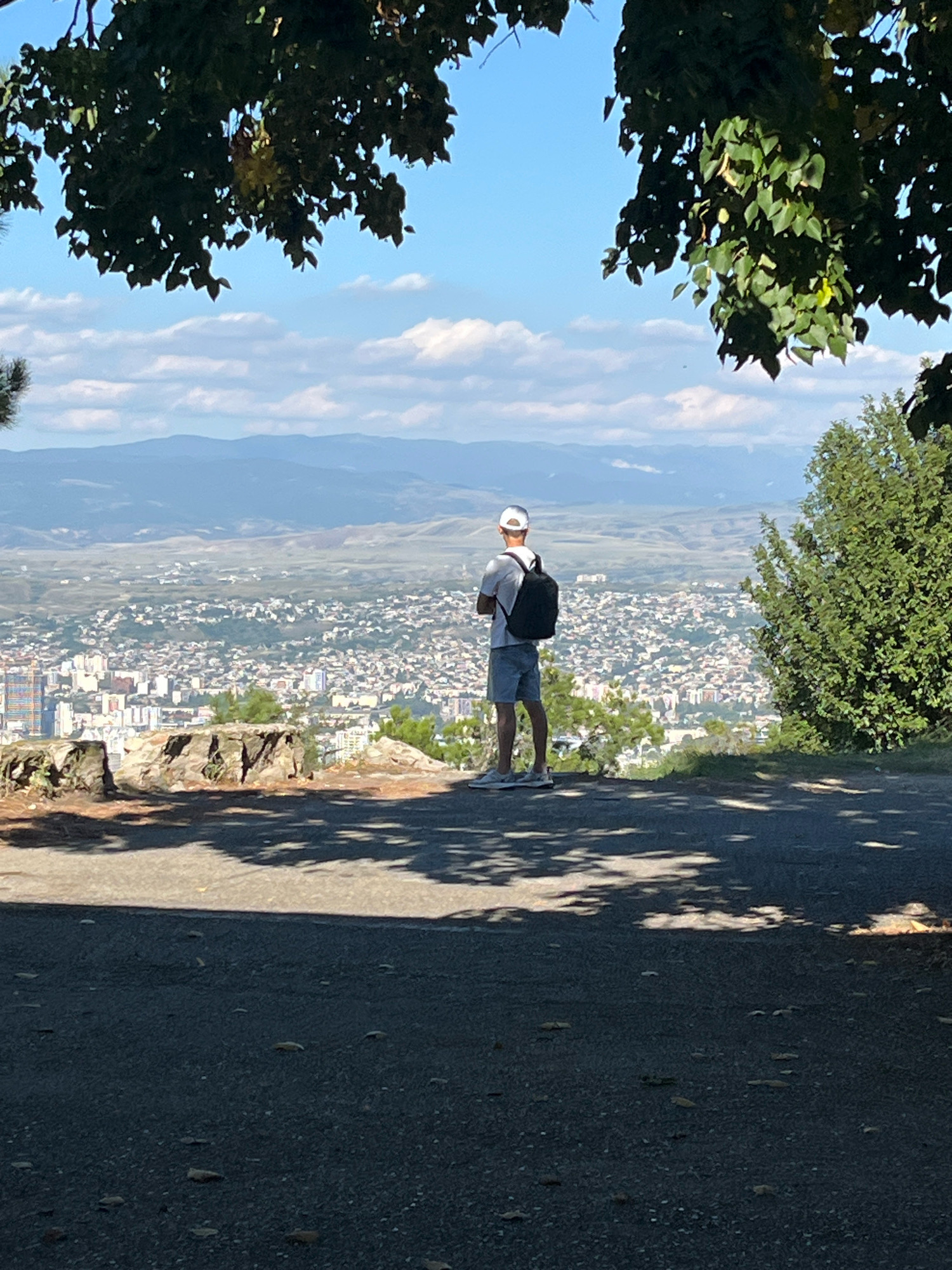 A lone individual stands on a paved path overlooking the sprawling city of Tbilisi, Georgia, from a high vantage point. The person, wearing a white cap, white t-shirt, and grey shorts, has a black backpack on their shoulders and is facing away from the camera, gazing at the cityscape. The sun casts dappled shadows on the path from the surrounding trees, some of which frame the top of the image. In the middle ground, a rocky outcrop is visible, and beyond that, the dense urban landscape of Tbilisi stretches out to the horizon, where distant mountains are faintly visible under a partly cloudy sky. To the right, a dense green tree adds to the natural elements of the foreground.