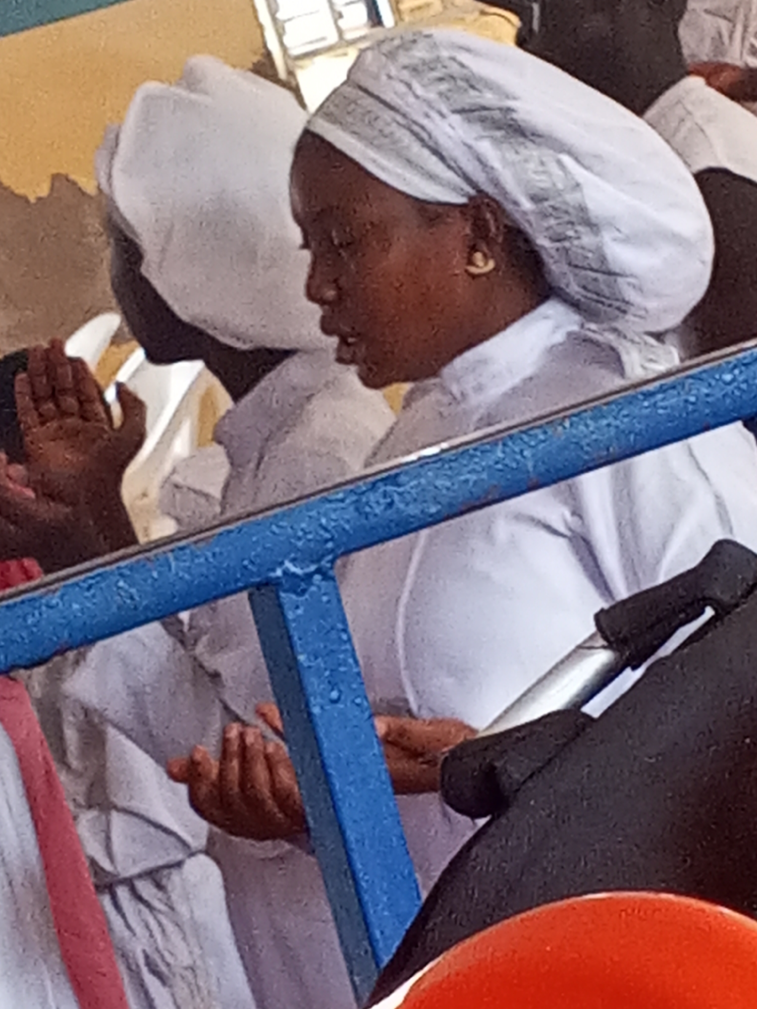 In Lokoja, Nigeria, several individuals are shown dressed in white robes and head coverings, suggesting a religious event or gathering. A woman with brown skin is in the foreground, wearing a white head covering and white robe. She has gold earrings and appears to be speaking or praying with hands clasped. Another person is next to her, also in white, with hands raised. A blue barrier separates the individuals from the viewer. A stroller is partially visible in the lower right corner. The background is indistinct, with hints of a painted yellow wall.