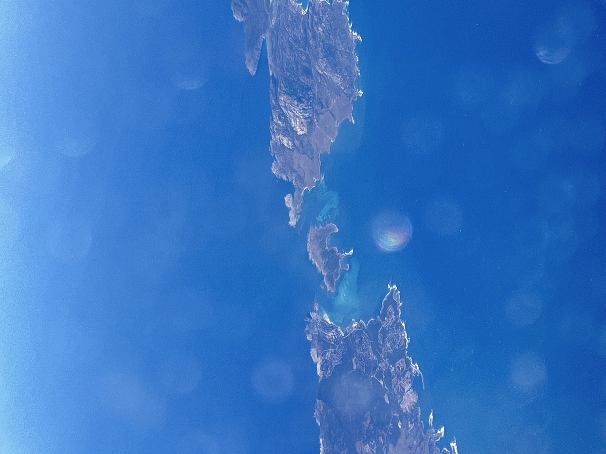 The image displays a high-altitude or satellite view of a long, narrow landmass, likely an island or a peninsula, surrounded by a vast expanse of bright blue water. The landmass stretches vertically through the frame, revealing a rugged, rocky, and somewhat barren terrain with irregular shorelines. Its surface shows textures and varying shades of grey and brown, consistent with mountainous or arid topography.
Around the landmass, especially in the shallower bays and channels, the water exhibits lighter, vibrant turquoise and teal tones, suggesting clear, shallow depths, sandy seabeds, or coral formations. The deeper surrounding ocean is a uniform, intense blue.
The scene appears to be taken under clear, sunny conditions, with no clouds visible. Several prominent circular and elliptical lens flares or bokeh artifacts are scattered across the blue water and sky areas, some displaying subtle rainbow hues, indicating the presence of a strong light source, likely the sun, and reflections within the camera lens. There are no visible human structures, activities, or discernible text within the frame. The image captures a pristine natural landscape from an elevated perspective.