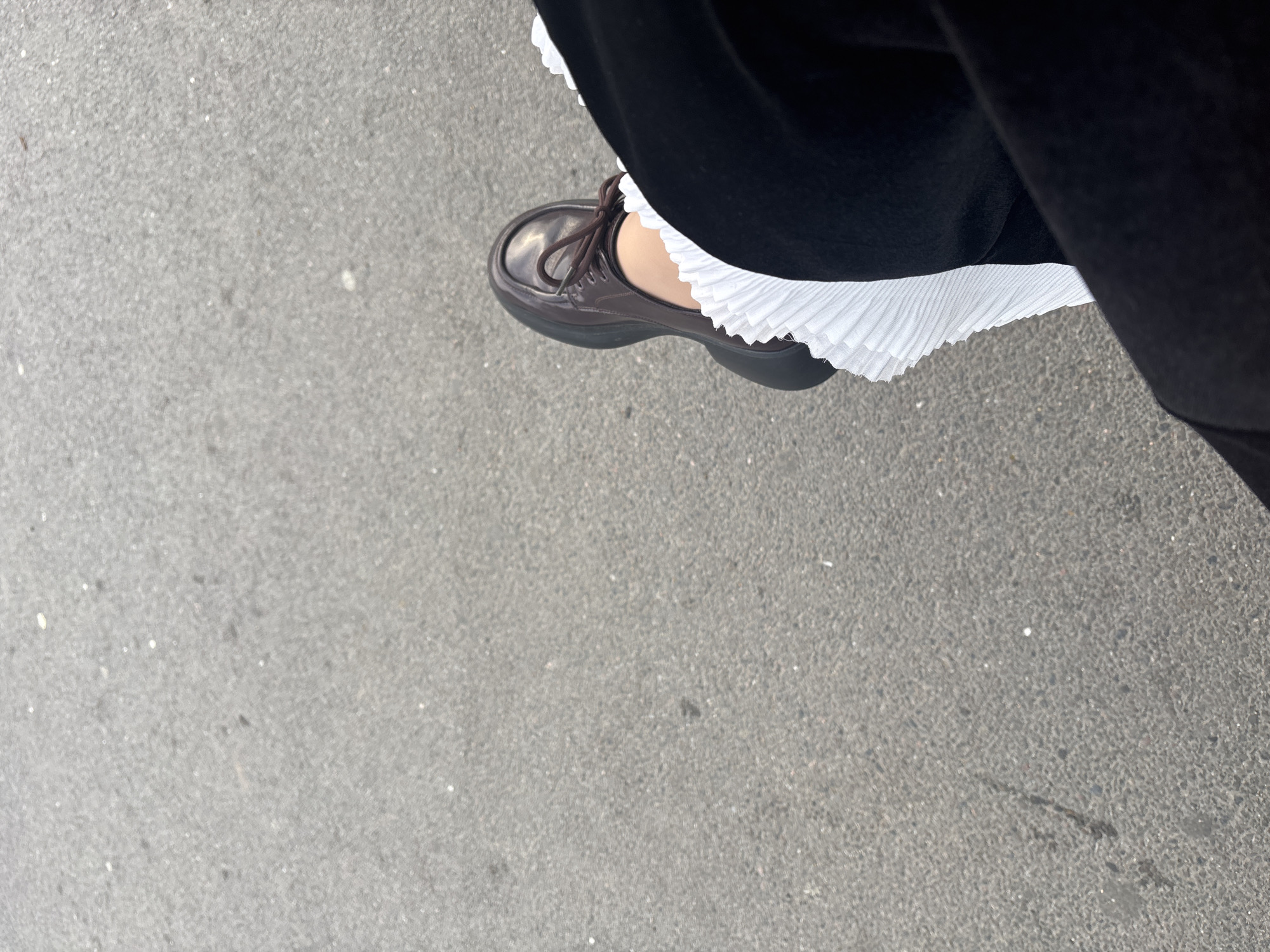 The image is a high-angle shot of a person's legs and shoes resting on a textured gray asphalt surface. The person is wearing dark pants, and a white pleated fabric, possibly part of a skirt or dress, is visible peeking out from under the pants and over a brown platform shoe. The shoe is laced and appears to be a derby or oxford style. The asphalt surface has small white specks and some faint dark marks. The image does not provide any specific visual cues to pinpoint its location within the city of Saint Petersburg, Russia, nor does it depict any interaction between individuals or specific actions beyond the resting posture of the person.